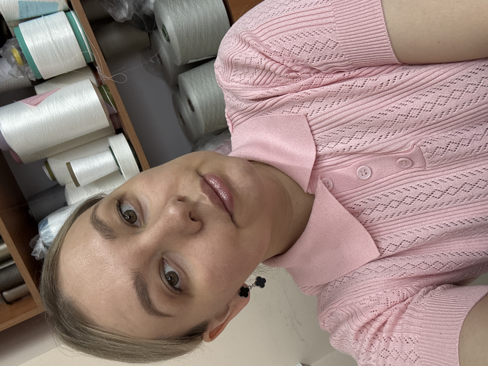 A selfie taken indoors shows a woman with light skin, brown eyes, and blonde hair, wearing a light pink, short-sleeved, button-down top with a collar, and flower-shaped earrings. She is looking at the camera. Behind her, there is a wooden shelf filled with spools of white yarn or thread. No information is available regarding the location in the city of Chaikovsky, Russia.