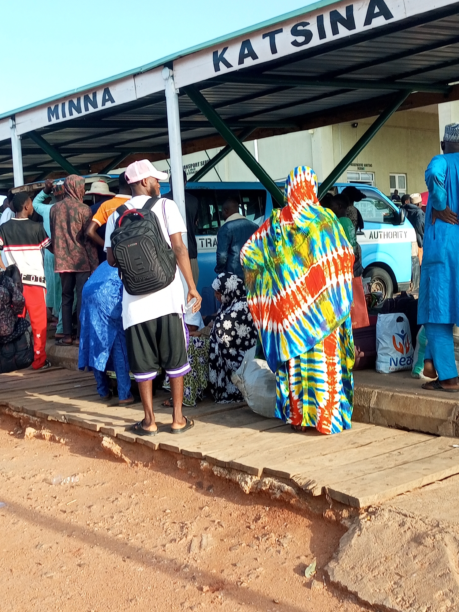 A group of people are gathered at a bus terminal in Gombe, Nigeria. Above them is a covered waiting area with signage that reads "MINNA" and "KATSINA". Several people are dressed in traditional Nigerian attire, including a woman in a brightly colored tie-dye dress and another in blue attire. Others are wearing more casual clothing, such as t-shirts, shorts, and pants. Some individuals are carrying bags, indicating they are likely travelers. A light blue bus with the word "AUTHORITY" on its side is parked nearby, suggesting it is a mode of transportation. The ground is made of reddish-brown dirt and a wooden platform is visible where the people are standing. The overall scene suggests a busy public transport hub.
