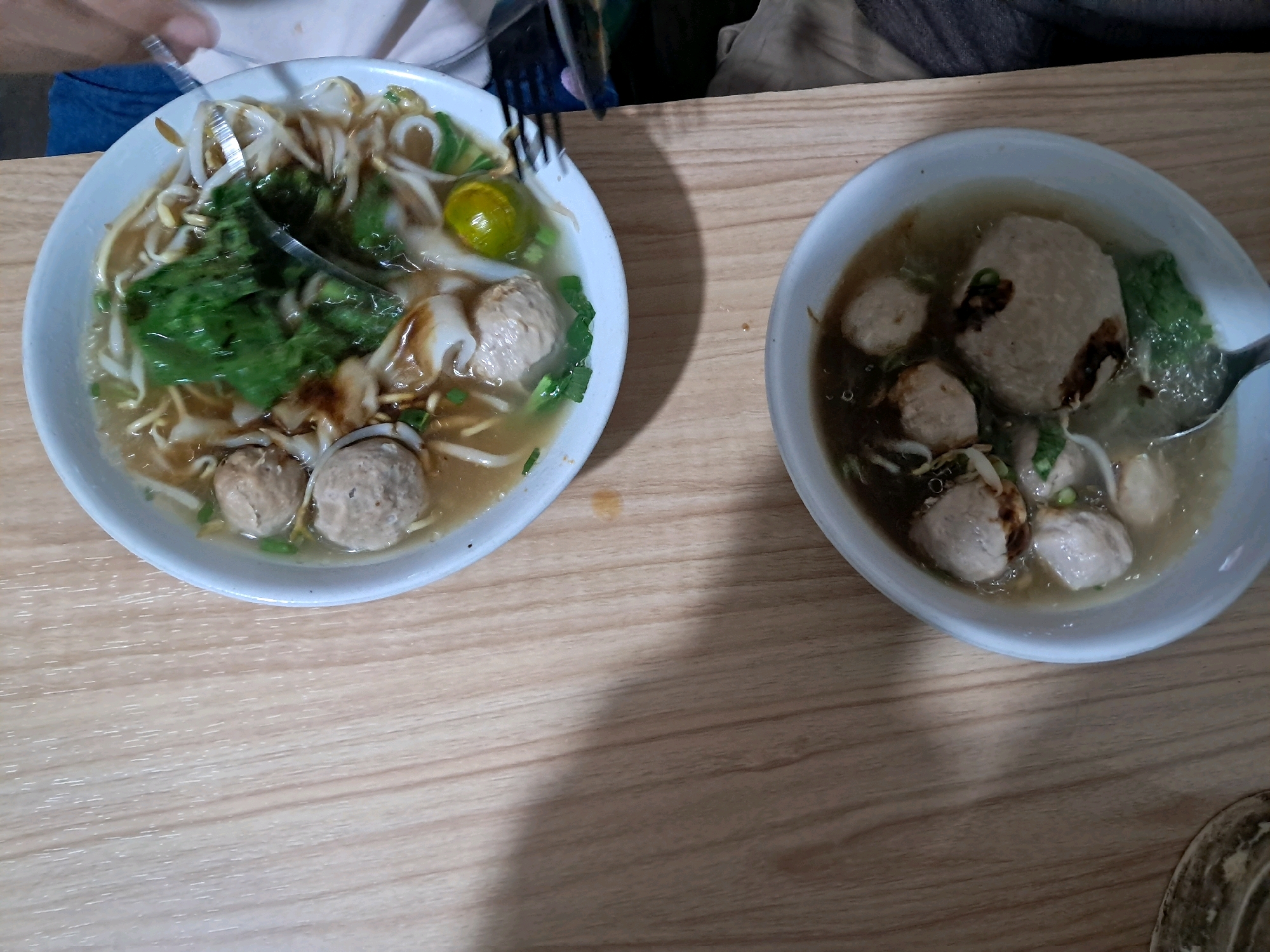 Two bowls of soup with meatballs are placed on a light-colored wooden table. The bowl on the left contains noodles, bean sprouts, leafy green vegetables, and several meatballs, with a slice of lime visible. A fork is being used to stir the contents of this bowl. The bowl on the right also contains a broth with several meatballs and some leafy green vegetables. The image is taken from a top-down perspective. The scene is set in Anjungan Melancar, Indonesia.