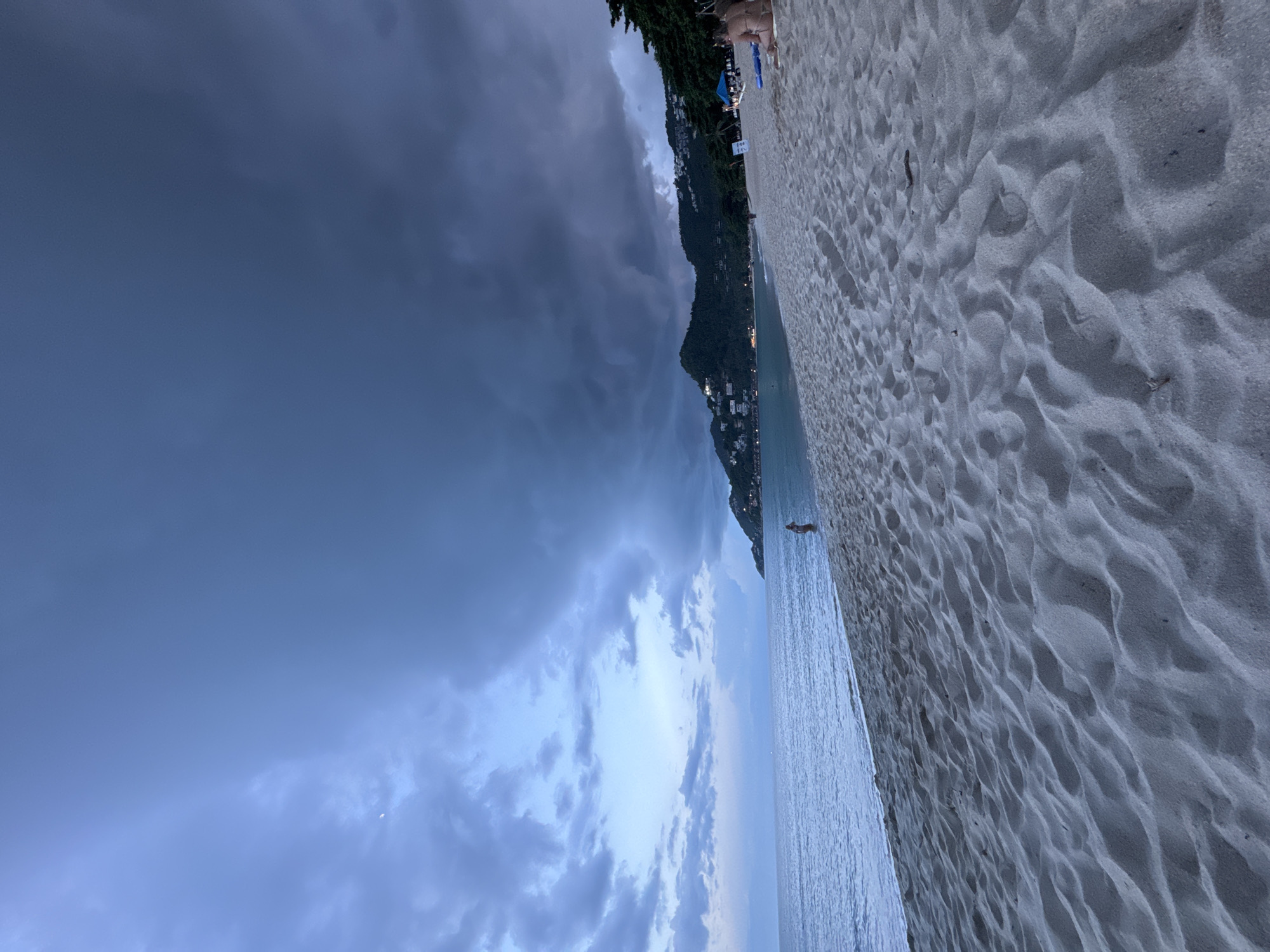 This media file depicts a beach scene at dusk. The sand on the beach is light-colored and textured with ripples from the wind or water. The ocean is calm with gentle waves lapping the shore. In the background, a lush green hillside is dotted with buildings, suggesting a coastal town or resort. The sky is dramatic with dark, ominous clouds gathering on the left side of the frame, contrasting with lighter, scattered clouds towards the horizon. The overall lighting suggests it is either early evening or a stormy pre-dawn hour. There are a few people visible: one person is lying on the sand towards the top right of the image, and another is walking near the water's edge on the right. There is also a small, dark shape that might be a person or animal further down the beach. A few beach chairs and umbrellas are also visible near the top of the frame. The mood is somewhat serene yet tinged with the drama of the approaching weather.