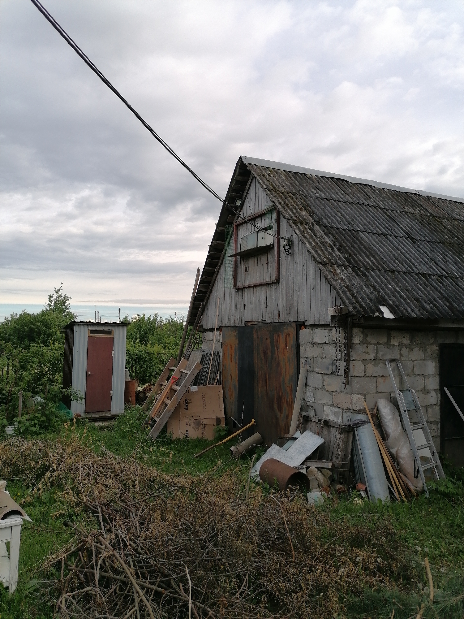 The scene depicts a cluttered backyard area with a shed and a small outhouse-like structure. The shed is constructed with a combination of materials, including wooden planks, cinder blocks, and a corrugated roof. An old window is visible near the top of the shed. Several ladders are leaning against the shed, along with various pieces of metal, wood, and debris. In front of the shed, a pile of dried branches and twigs is scattered on the grass. The outhouse-like structure has a simple design with a red door. The sky is overcast with clouds, and in the distance, a glimpse of a body of water can be seen. The location is an unidentified area with vegetation and unkempt surroundings. No people are visible in the scene.