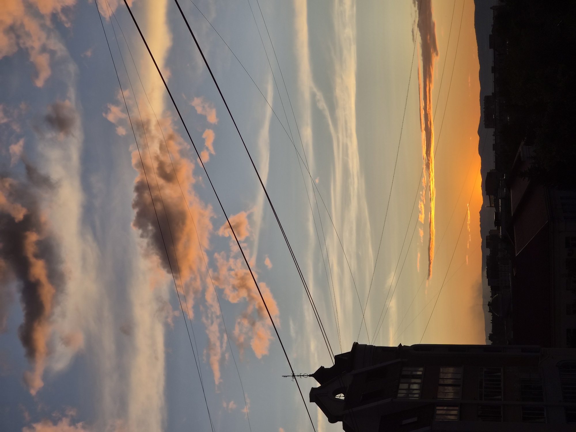 The image depicts a sunset over the city of Chita, Russia. In the foreground, a silhouette of a building with a pointed roof and architectural details is visible on the left. The sky is filled with colorful clouds, ranging from pale orange and yellow near the horizon to darker blues and grays higher up. Several power lines crisscross the sky. In the background, a silhouetted cityscape with mountains beyond is dimly visible, with the sun appearing as a bright orange glow on the horizon. The overall scene conveys a tranquil, late-evening atmosphere.