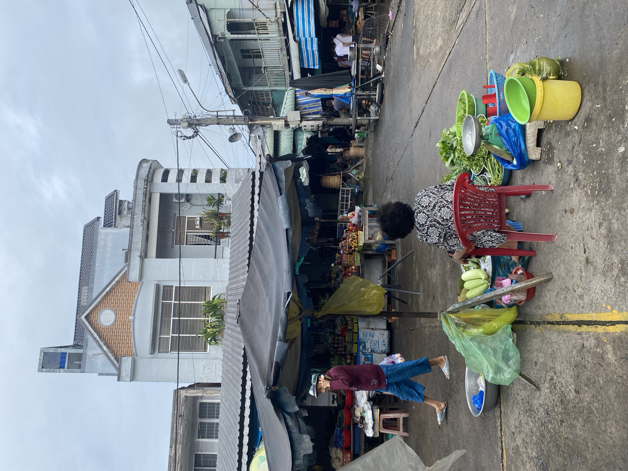 An outdoor market scene is depicted under an overcast sky. In the foreground, a woman with dark, curly hair sits on a red plastic chair facing away from the camera, wearing a patterned black and white shirt. In front of her are various vegetables in bowls, including leafy greens and what appear to be gourds or cucumbers, a green kitchen scale, and colorful plastic containers. A yellow bucket and a large green plastic bag are also visible. To the left, a person in blue pants and a burgundy top walks away from the camera, holding a plastic bag. They are near a metal basin and a stool. The background features market stalls with awnings and displays of goods, including food items and what appear to be bottled products. Buildings of varying architectural styles are visible behind the market stalls, including a multi-story structure with a distinctive roofline. Electrical wires and a utility pole are prominent features in the background. The ground is paved and wet in some areas.