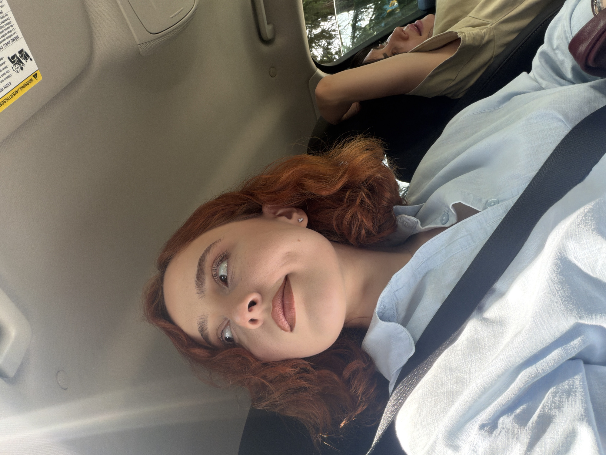 This is an eye-level, indoor shot of two people in a car. The first person, a young woman with red, wavy hair, fair skin, and brown lipstick, is looking at the camera. She is wearing a light blue shirt and a seatbelt. The second person is lying down in the background and is wearing a beige shirt. The background appears to show trees and sunlight. The setting appears to be inside a car. There is no indication of the location being Tbilisi, Georgia.