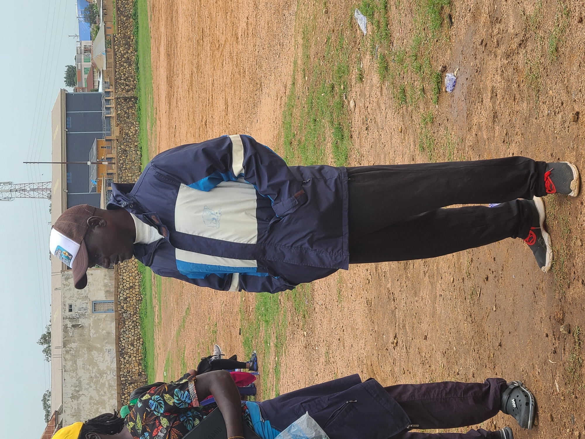 The image shows a man in Unknown, Unknown, wearing a brown and white baseball cap, a blue and white jacket, and black pants. He stands on a dirt and grass ground. In the background are buildings, a stone wall, and an electrical tower. Another person is partially visible at the bottom of the image wearing a floral shirt and black pants.