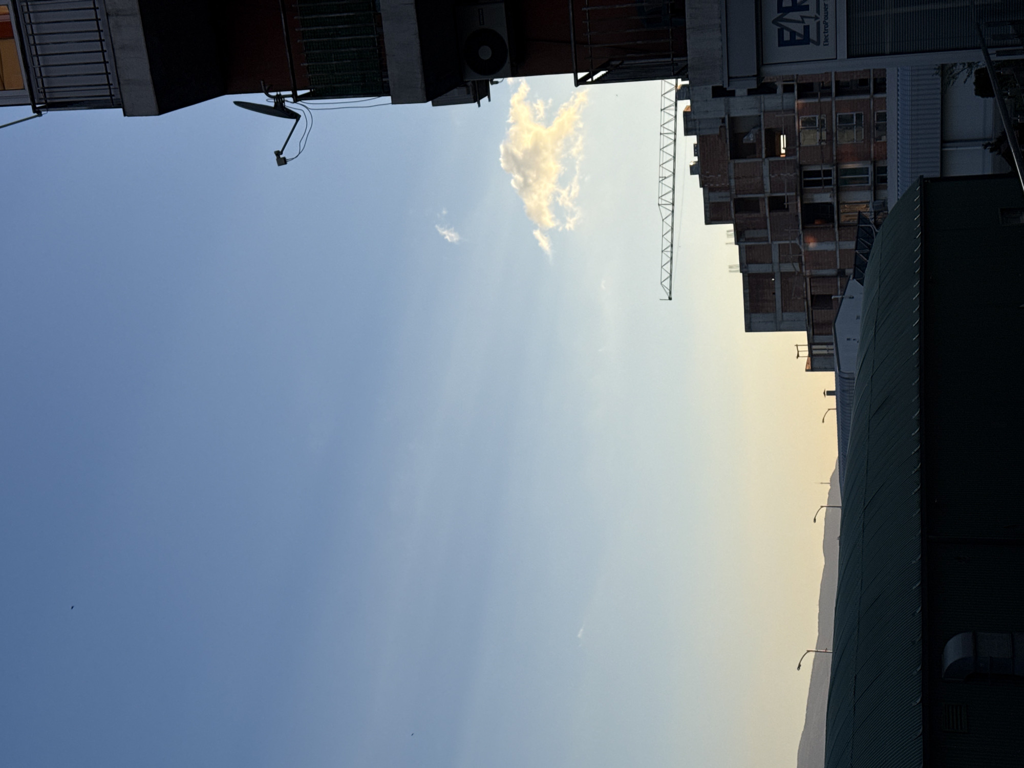 The image depicts an urban scene in Sofia, Bulgaria, focusing on the sky framed by buildings. The sky transitions from a light blue to a yellowish hue near the horizon, with a few scattered clouds. A construction crane is visible near a building under construction. There are residential buildings on the left and a large industrial building to the right. A satellite dish is mounted on one of the buildings.