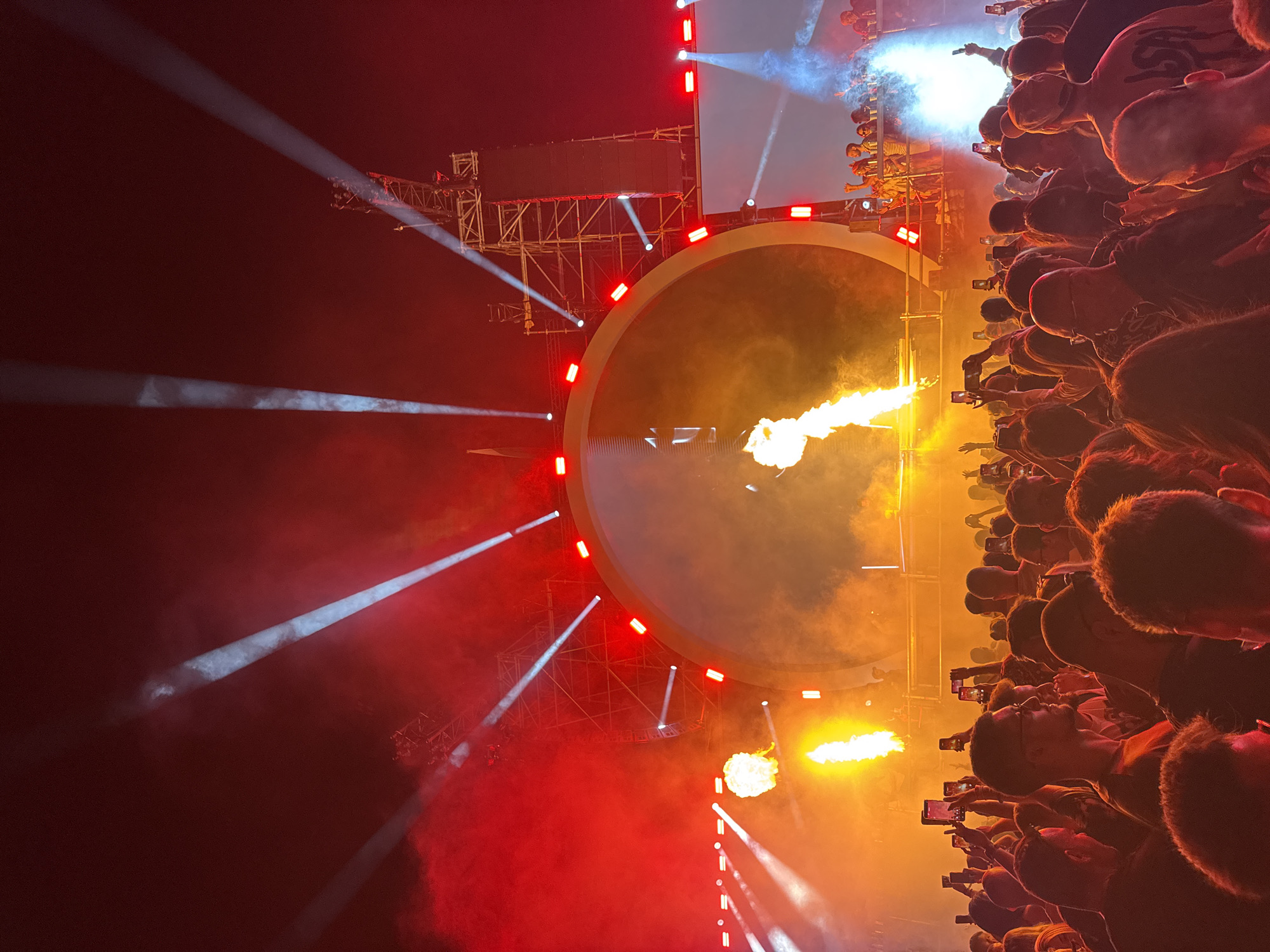 The image depicts a large outdoor concert or festival in Sofia, Bulgaria. The foreground is filled with a densely packed crowd of people, many holding up smartphones to record or photograph the event. The crowd is illuminated by a vibrant red and orange stage lighting, with plumes of smoke and dramatic flame effects rising from the stage.
The stage itself is a prominent feature, dominated by a large, circular screen or LED display with red lighting around its circumference. Two large, intense flame bursts are visible emanating from the lower part of the stage. Above the stage, a complex metal truss structure supports lighting rigs and large speakers. White spotlights cut through the smoky atmosphere, creating beams of light that extend upwards into the dark sky. A large screen or projection surface is visible to the right of the main stage. Several individuals are visible on an elevated platform or walkway at the rear of the stage, seemingly performing or interacting with the crowd. The overall atmosphere is energetic and immersive, characteristic of a large-scale music event.