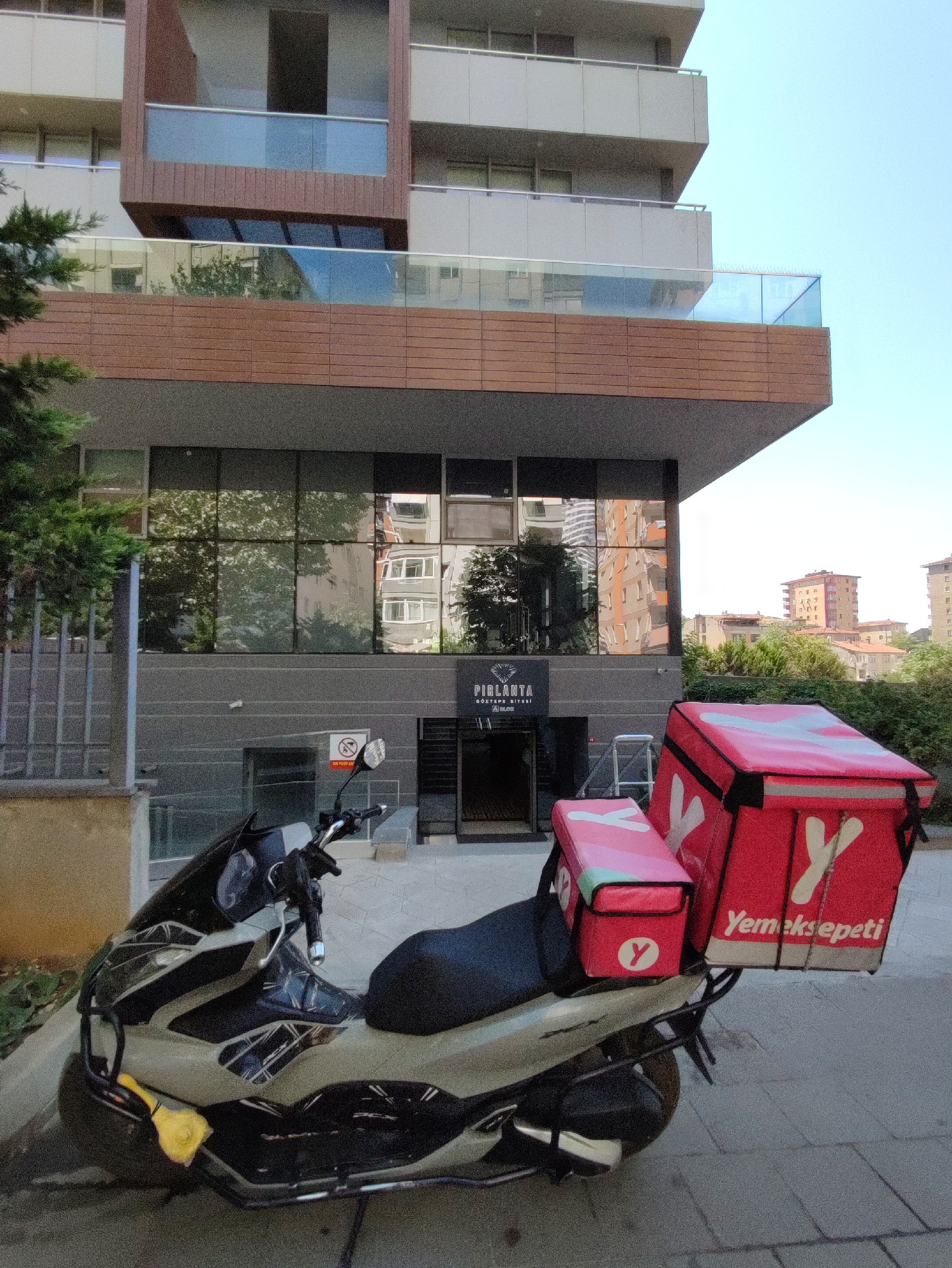 The image depicts a scooter parked in front of a modern building with a glass facade and balconies. The scooter is equipped with two red delivery bags, one larger on the rear and a smaller one on the front. Both bags feature the "Yemeksepeti" logo, indicating a food delivery service. A sign on the building reads "PIRLANTA GÖZTEPE SITESI," suggesting the location is Pirlanta Göztepe Sitesi in Kadıköy, Turkey. The building appears to be an apartment complex or a commercial building. Reflections in the glass facade show trees and other buildings. The sky is clear and blue. No people are visible in the image.