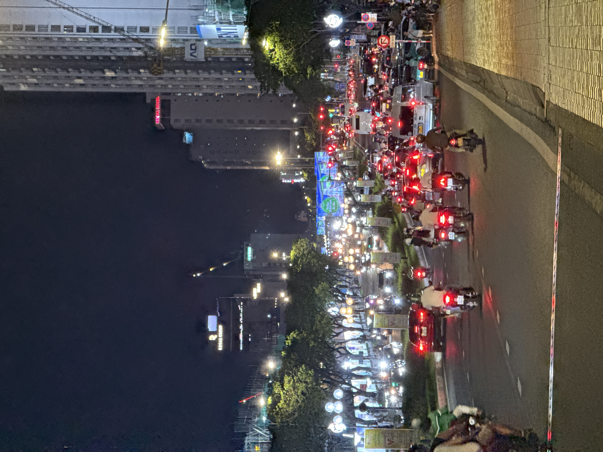 This image captures a vibrant and busy street scene at night in Đà Nẵng, Vietnam. The time of day is clearly evening or night, indicated by the dark sky and the numerous artificial light sources illuminating the street and buildings.
The primary subjects are the dense flow of traffic, overwhelmingly composed of motorbikes, with some cars interspersed. Hundreds of bright red taillights from vehicles dominate the foreground and midground, suggesting significant congestion or a stop at a traffic light. Many individuals on motorbikes, some wearing helmets, are visible. The activity taking place is typical urban evening commuting, with vehicles either stopped or moving slowly along a multi-lane road.
On the left side of the street, a series of tall, multi-story buildings rise into the dark. One building features a bright red neon sign that clearly reads "GERAVA HOTEL." Further down, another illuminated sign displays "BROAD GROUP." A large construction crane is also visible near one of the taller buildings, indicating ongoing development. Streetlights and trees, some adorned with decorative lights, line the thoroughfare. Traffic signs, including a red circular "no entry" symbol and an arrow sign, are visible above the road. On the right, a textured, light-colored wall or barrier runs along the side of the road, separating it from an unseen area. The overall scene conveys the bustling energy of a city at night.