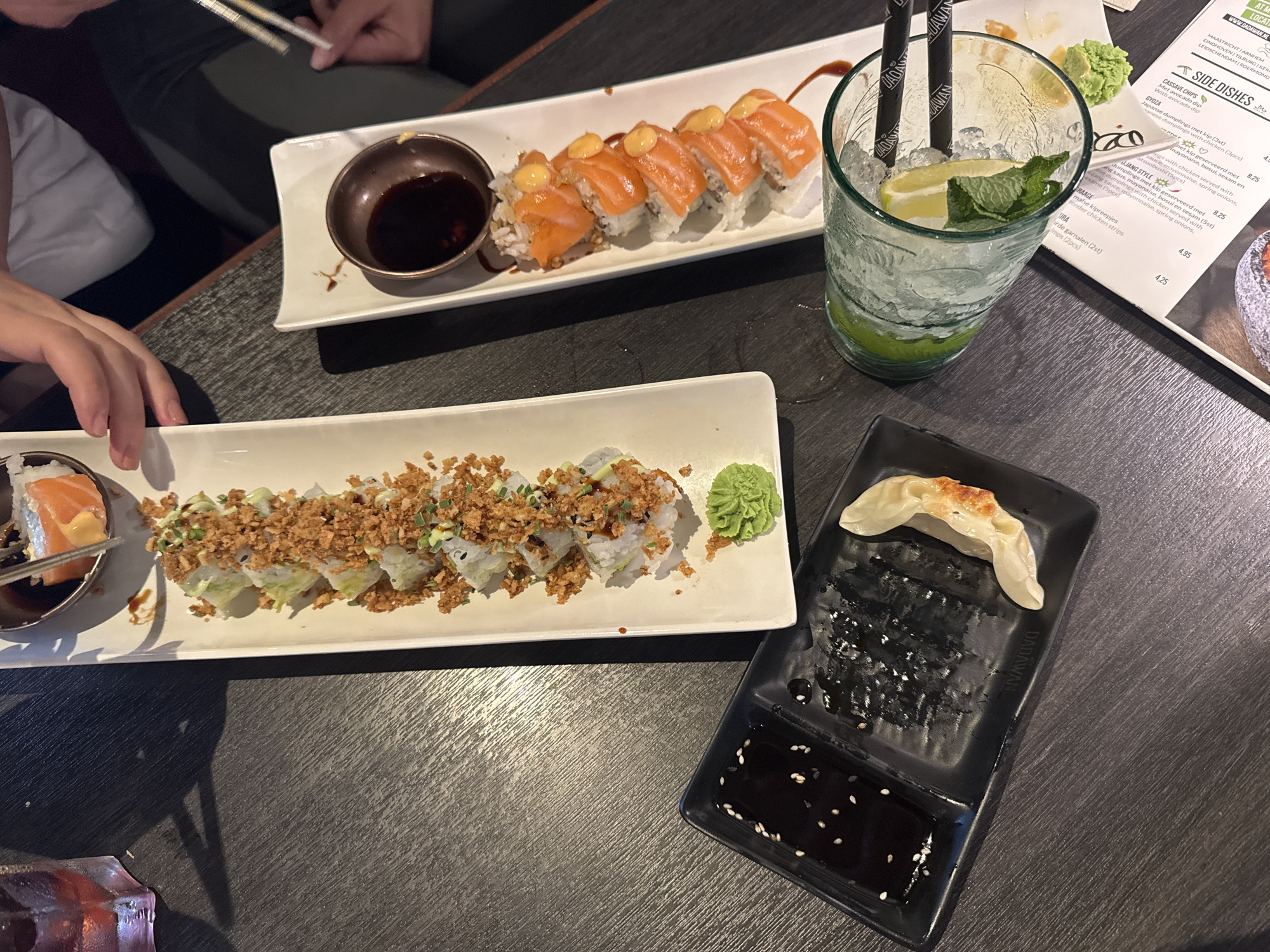 The image depicts a close-up, overhead view of a table at an Asian restaurant, likely in Leidschendam, Netherlands. The table is dark and wooden with a visible grain. On the table are two plates of sushi: one contains several pieces of sushi topped with crispy fried onions and green onions, and another plate features sushi rolls topped with salmon and a creamy sauce. Adjacent to the sushi is a small dish with soy sauce and a dollop of green wasabi. A black rectangular plate holds soy sauce in one compartment and a single, golden-brown gyoza in another, with some dark, shredded material. To the right, a tall, clear glass is filled with ice, mint leaves, and lime wedges, with two black chopsticks resting inside. A menu is partially visible at the bottom right. Two pairs of hands are visible, one holding chopsticks and reaching for sushi, and another partially obscuring a white shirt. The lighting suggests an indoor setting.