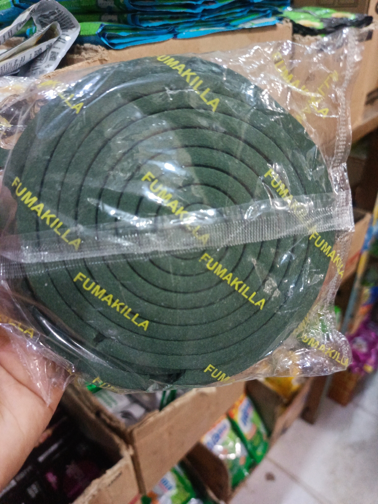 The image shows a roll of green mosquito coils packaged in clear plastic. The brand name "FUMAKILLA" is printed in yellow on the plastic packaging, repeating around the coil. The mosquito coil is dark green and appears to be made of a tightly wound material. The packaging is slightly creased and reflects light in some areas. The mosquito coil is positioned in the foreground, taking up a significant portion of the frame. In the background, a hand is partially visible on the left, holding the packaging. Behind the mosquito coil, there are blurred images of other products in cardboard boxes and on shelves, suggesting a retail or market environment. The background also includes glimpses of colorful packaging, possibly of other household or personal care items. The overall impression is of a product displayed for sale. The location is identified as Ilotidea, Indonesia.