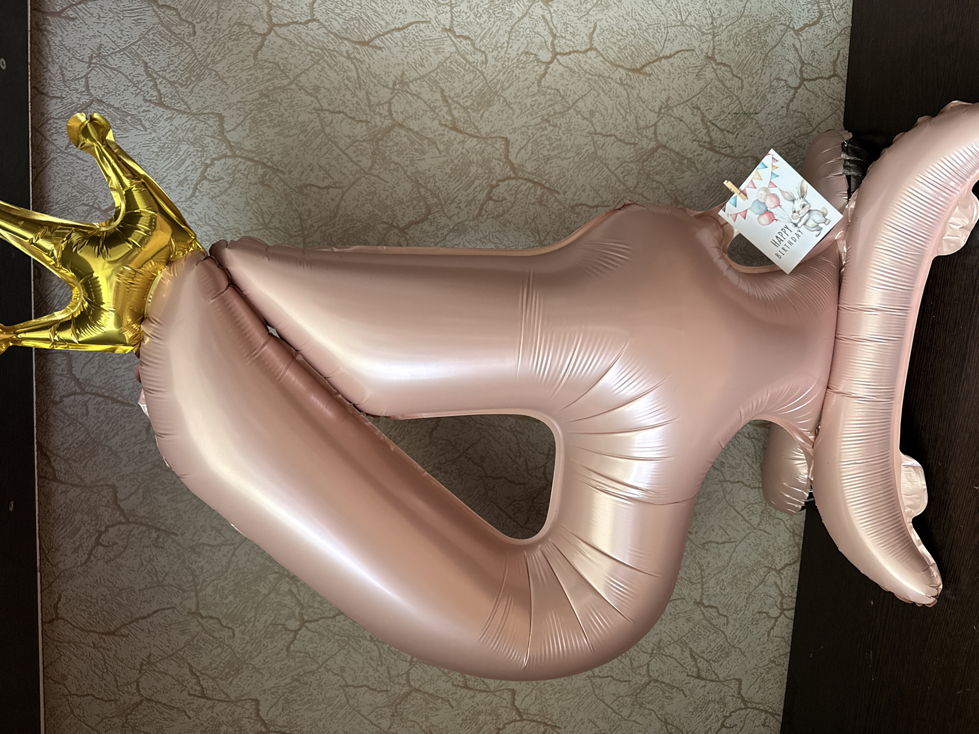 A large, rose gold foil balloon shaped like the number '4' stands upright, topped with a shiny gold crown balloon. A small, square card, clipped to the side of the '4' balloon with a miniature clothespin, features a watercolor illustration of a rabbit holding balloons and the text "HAPPY BIRTHDAY". The balloons are positioned on a dark, glossy surface, possibly a table, against a textured, light brown wallpaper. The scene is indoors and the lighting suggests an ambient indoor environment. There is no information to indicate the location is Mezhdurechensk, Russia.