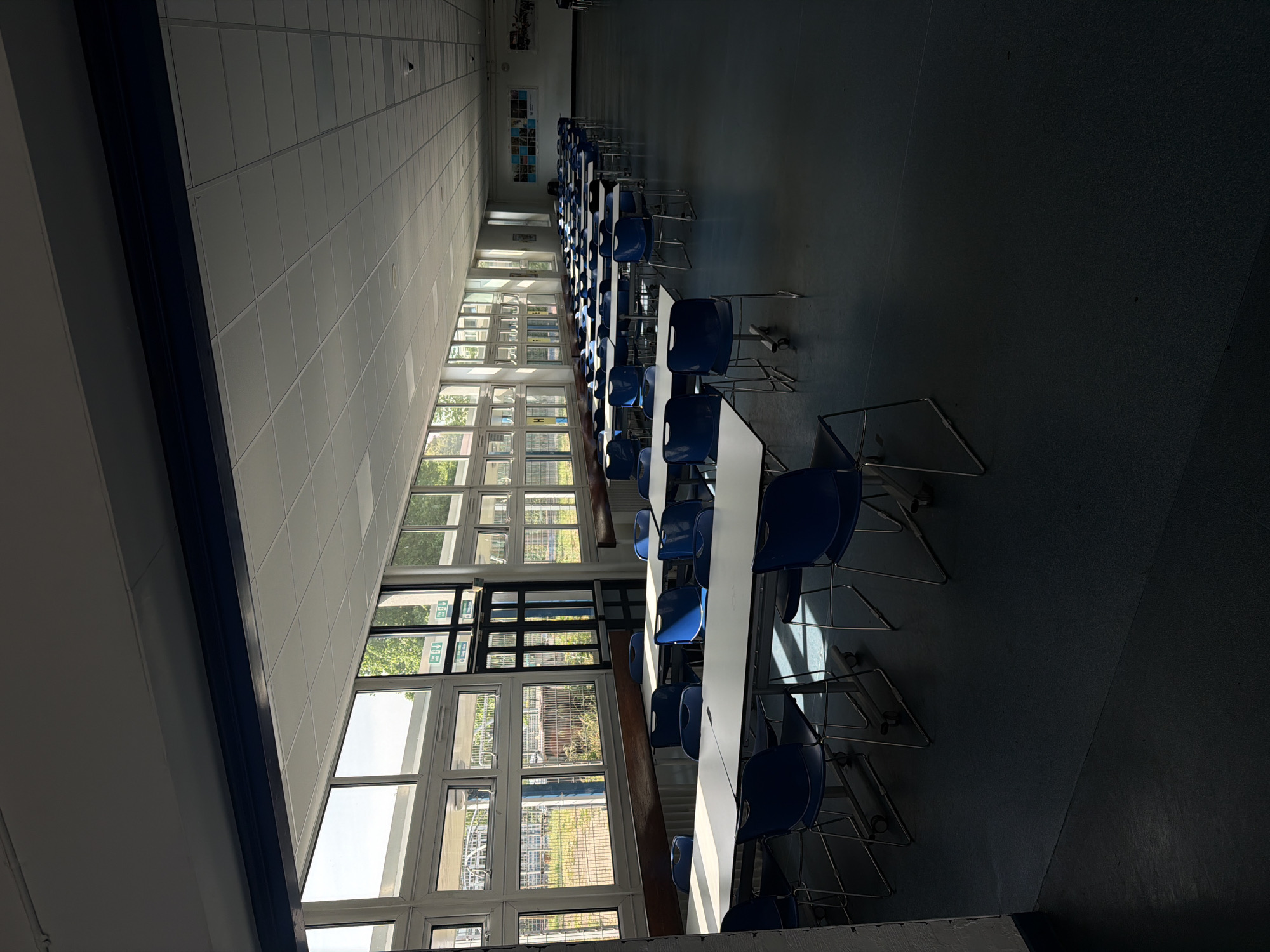 The image depicts a long, empty dining hall or cafeteria. Rows of white tables with blue chairs are aligned in neat rows, stretching into the distance. Large windows along one side of the hall allow natural light to stream in, creating strong patterns of light and shadow on the floor and furniture. An exit sign can be seen near the windows. The walls and ceiling are predominantly white. There is no indication of the location being in London, United Kingdom. No people are visible.