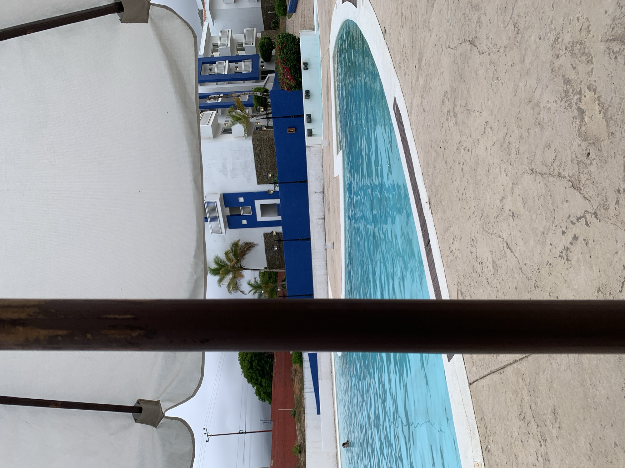 The image presents an outdoor scene in Playa El Agua, Venezuela, viewed from under a white patio umbrella. A dark brown, likely metal or painted wooden, umbrella pole occupies the central foreground, extending from top to bottom. The umbrella's white fabric is visible at the top and sides of the frame. The ground consists of light beige, textured stamped concrete, exhibiting cracks and minor discoloration.
In the midground, a light blue swimming pool with visible surface ripples features white coping and a dark trim along its edge. A single individual with dark hair is partially submerged in the left section of the pool, with their head and shoulders visible above the water, indicating swimming activity.
Beyond the pool, a low white retaining wall precedes a distinct dark blue wall or fence. Several black lampposts with dark fixtures are regularly spaced along this blue barrier. The background features multiple multi-story buildings characterized by white facades and prominent blue architectural elements, including recessed sections and balconies with white railings. Green foliage, including several palm trees and a bush with red blossoms, is distributed throughout the background. To the far left, utility poles and power lines are visible against a uniformly overcast grey sky, situated above a red earth bank covered with dense green trees.