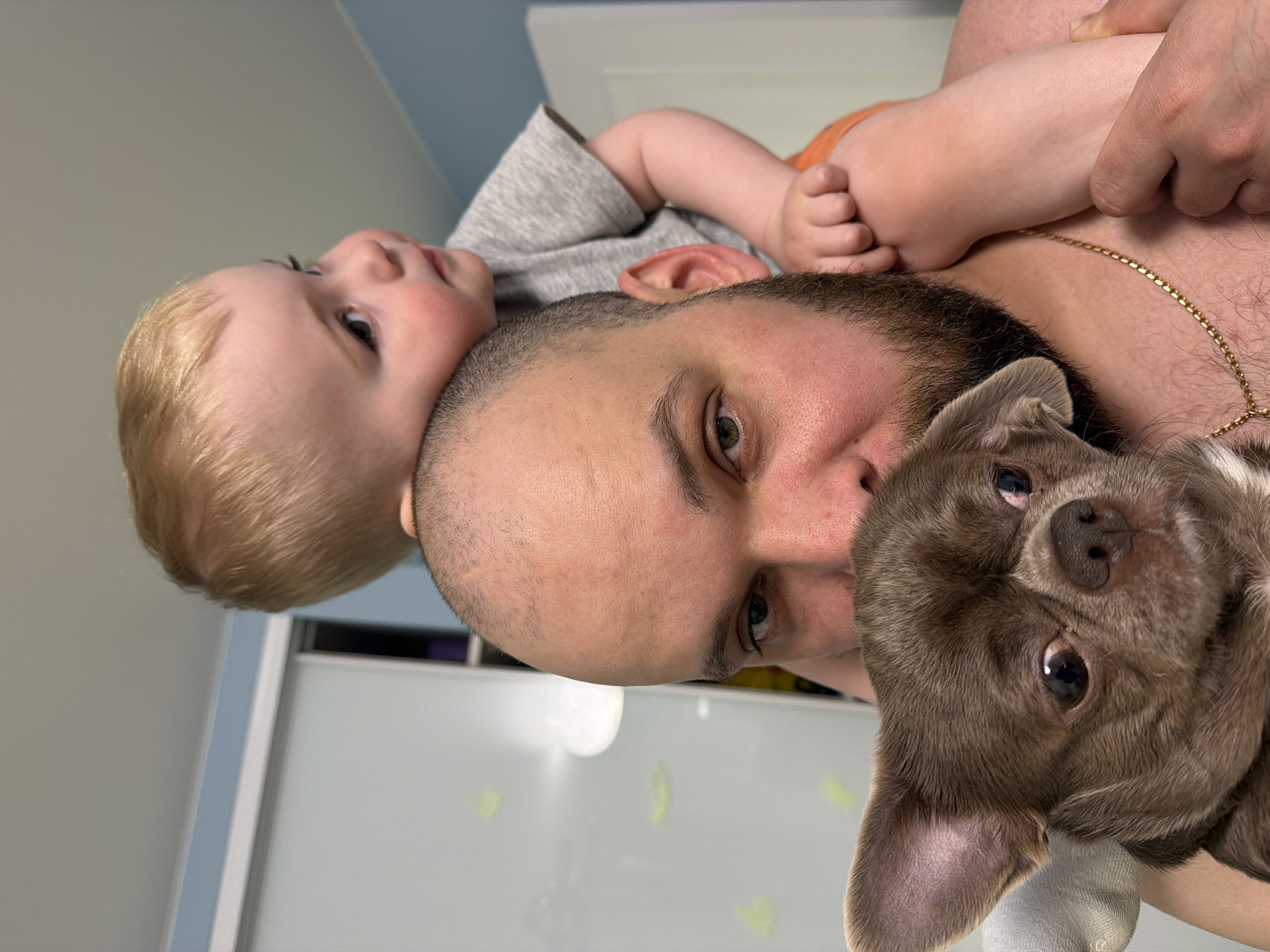 A photo of a man, a baby, and a dog. The man is balding with a beard and is facing the camera. The baby is lying on the man's shoulder and has blonde hair. The dog, a Chihuahua, is being held in the man's arms. The photo was taken indoors, with a window and white wall in the background. There is no information to determine the specific location in Votkinsk, Russia, or any other city.