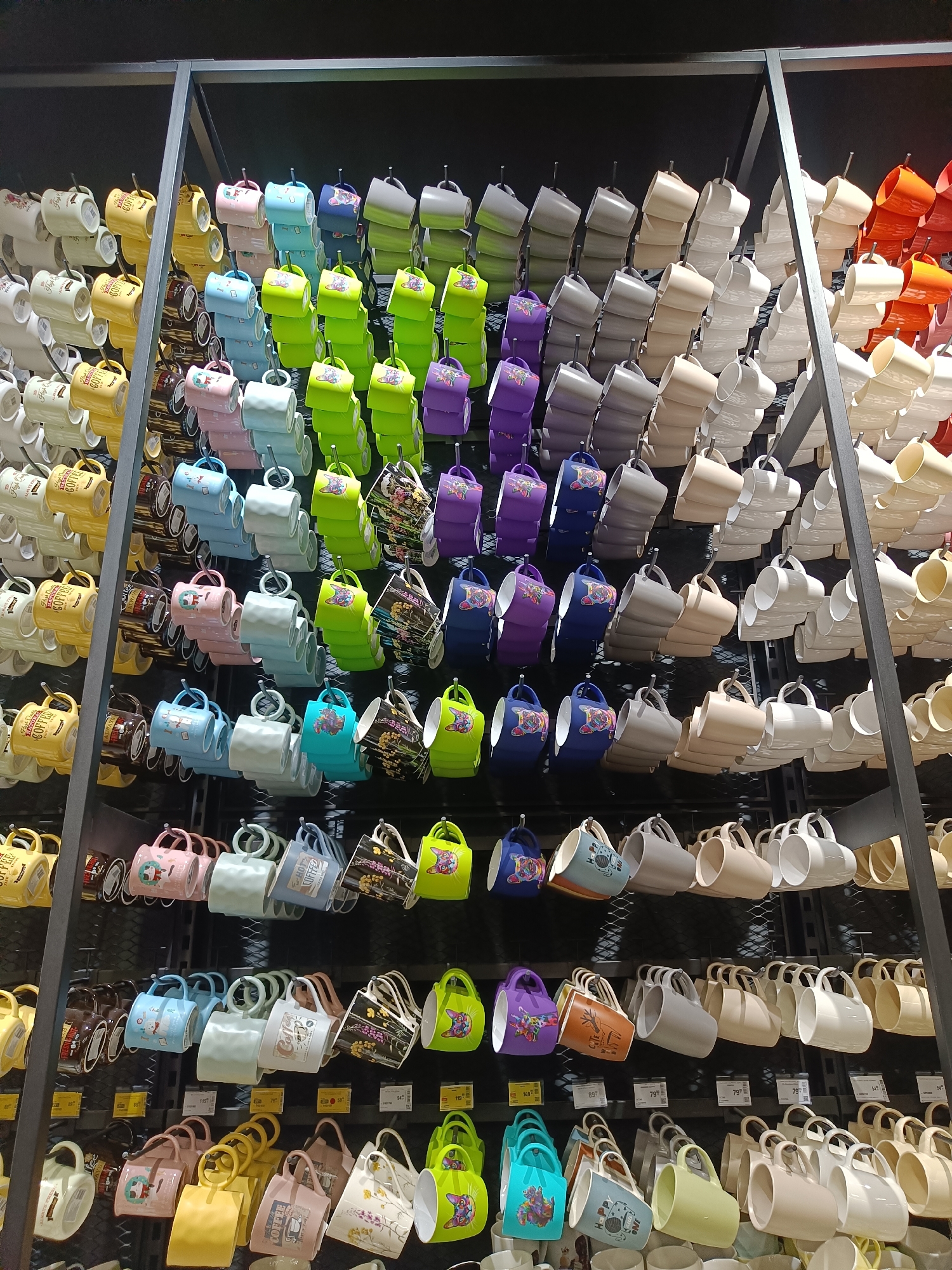 The image shows a retail display of mugs in a store. The mugs are arranged on multiple shelves, hanging from hooks. There are a variety of colors and designs, including solid colors, patterned mugs, and mugs with cartoon characters. The mugs are made of ceramic or a similar material. The display is well-lit, and the background is dark, which makes the colorful mugs stand out. There are no people visible in the image, and no actions are taking place other than the display of merchandise. The store appears to be a general merchandise store or a gift shop. There is no indication of the location being Vinnytsia, Ukraine.