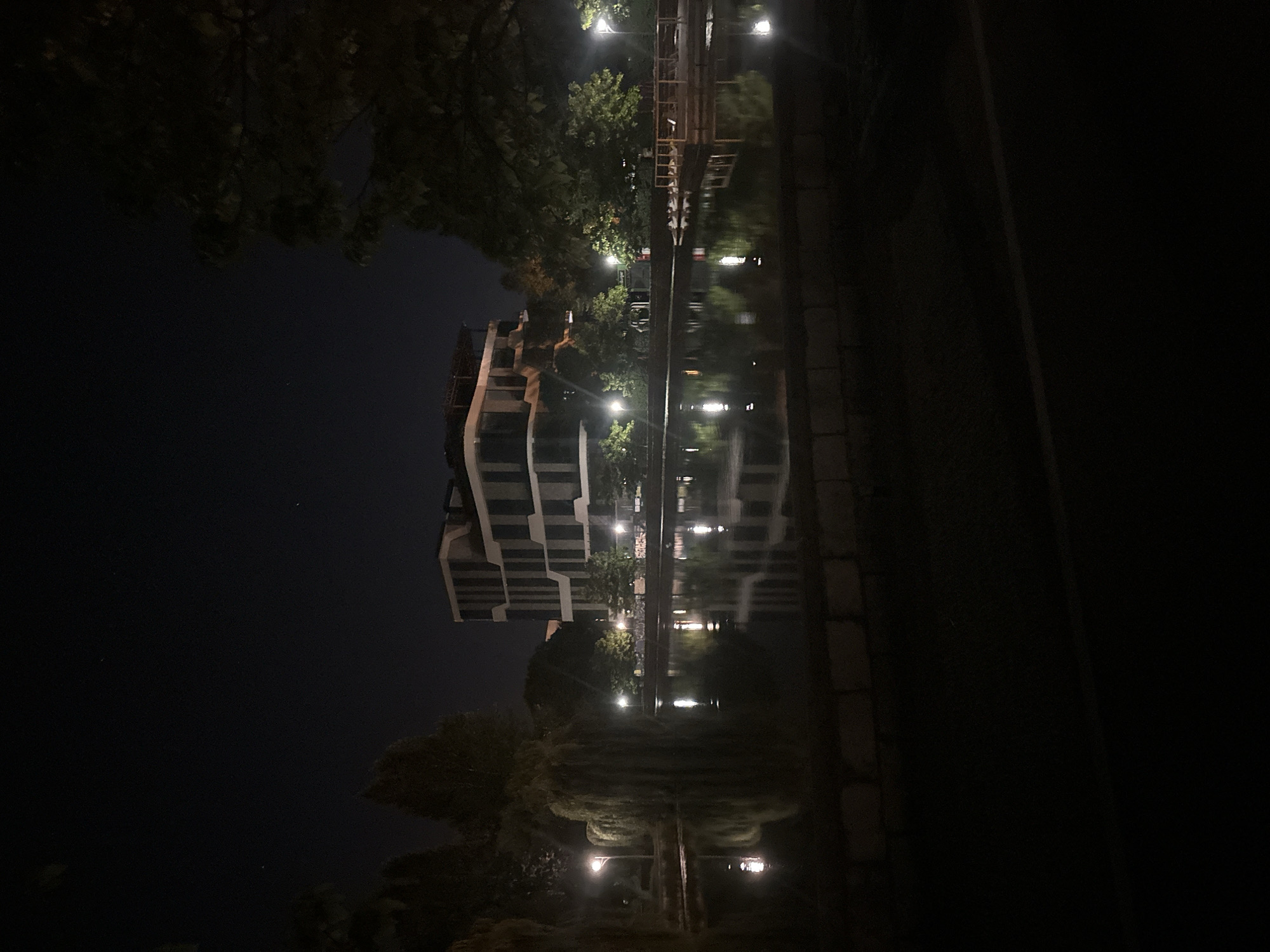 The image depicts a nighttime scene in Ternopil, Ukraine, featuring a tranquil pond reflecting the lights of a modern building and streetlamps. The building, with its distinctive geometric design and dark facade, stands prominently in the background, partially obscured by trees. The pond's surface is calm, creating clear reflections of the surrounding illumination.
The environment suggests a park or public space, evidenced by the presence of streetlights and the arrangement of trees. One particularly noticeable tree with drooping branches, likely a weeping willow, adds a touch of natural beauty to the urban landscape. The sky is dark, indicating late evening or early morning, with a few faint stars visible. The foreground is dominated by darkness, with a hint of a paved surface and a low brick wall, suggesting a pathway or embankment. No people or animals are visible in the frame. The overall atmosphere is peaceful and serene, highlighting the interplay of light and shadow in an urban setting.
