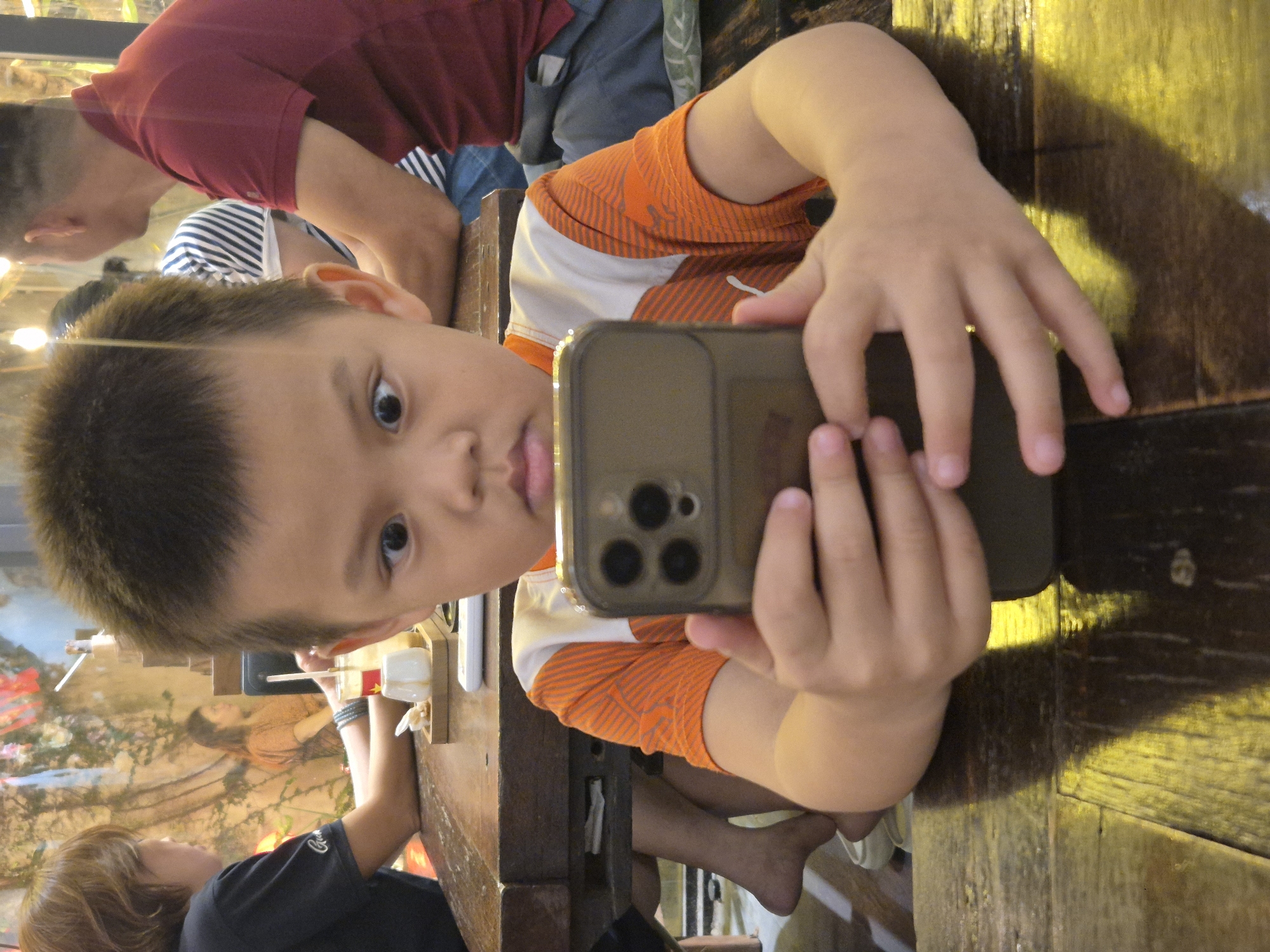 A young boy is sitting at a wooden table, holding a smartphone and looking at it intently. He is wearing an orange and white striped t-shirt. His eyes are wide open and looking slightly to the left. In the background, there are other people sitting at tables. The environment appears to be an indoor restaurant or cafe with a mural on the wall. The lighting is warm and there are reflections on the table. The specific location in Da Nang, Vietnam is not discernible from the image.