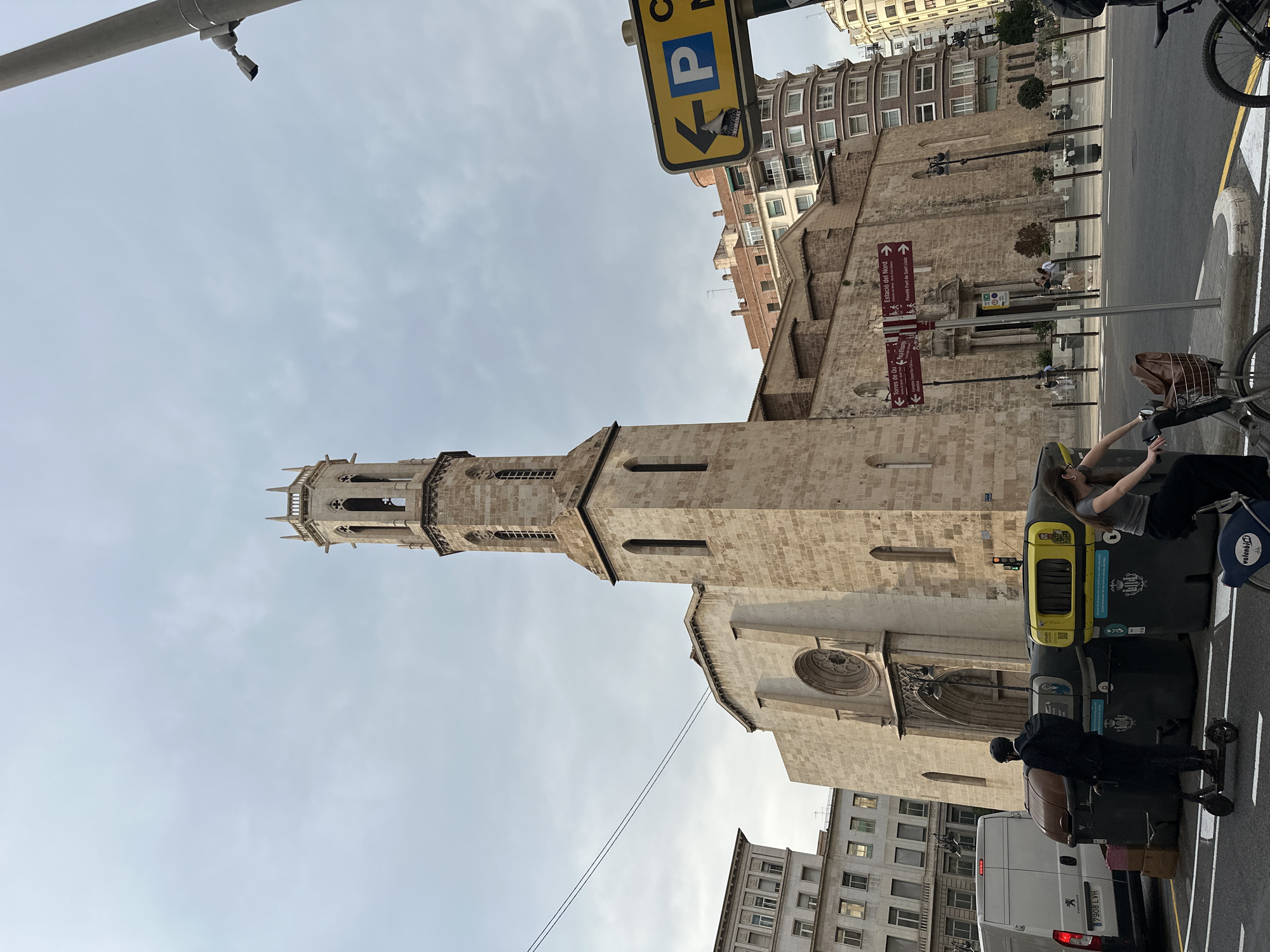 The image depicts the Church of San Nicolás de Bari y San Pedro Mártir in València, Spain, a historic religious building constructed primarily of beige stone. The church features a prominent bell tower with Gothic architectural elements, including arched openings and decorative spires. Adjacent to the church are other urban buildings, including a multi-story building with many windows and a curved, modern-looking building with balconies.
In the foreground, a street scene unfolds. A white van with a license plate reading "7908 LVH" is parked on the left. Beside it are several large waste bins, two of which are dark colored and one is yellow. A person wearing a black helmet and dark clothing is standing on a black electric scooter, facing away from the camera. To the right of the waste bins, a person is riding a bicycle and looking to their right.
A yellow directional sign with a blue "P" symbol indicating parking is visible on a pole on the right side of the image. Below it, a red directional sign points left towards "Torres de Quart" and "Campus d'Estudi General" and right towards "Estació del Nord" and "Pavelló Font de Sant Lluís". The sky is overcast with a cloudy, light gray appearance. The overall impression is an urban street scene with historical architecture.