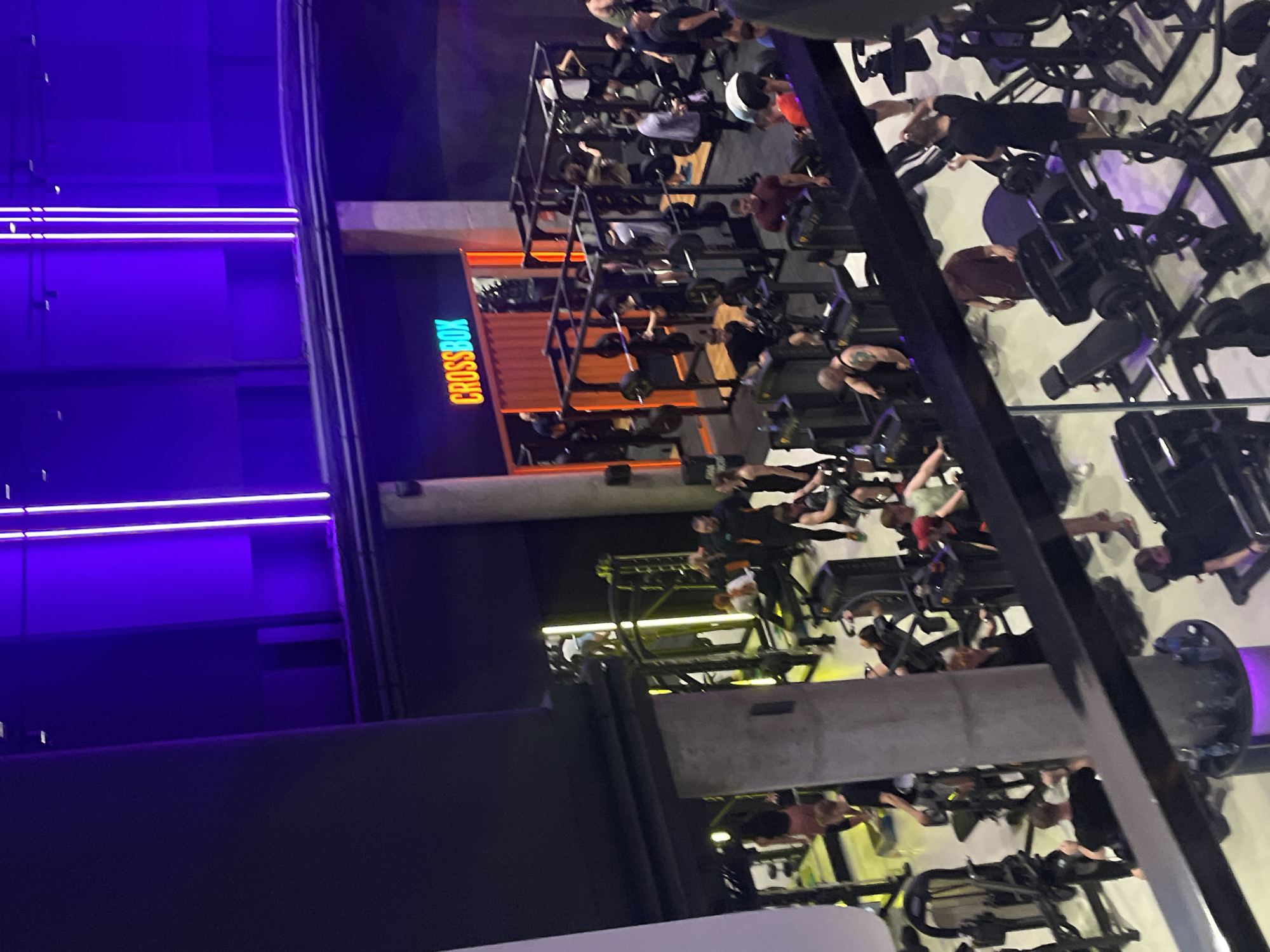 The image shows the interior of a fitness center in Moscow, Russia, specifically a "Crossbox" facility. The scene includes multiple levels with exercise equipment and people engaged in various workout activities. The environment features a modern, industrial aesthetic with bright lighting, including neon or LED lighting with a blue-violet spectrum, as well as orange accents on the Crossbox branding. The facility appears to be busy with a diverse group of individuals. A variety of fitness machines can be seen, with people actively using them. The layout is multi-tiered, showcasing a multi-level approach to the fitness space.