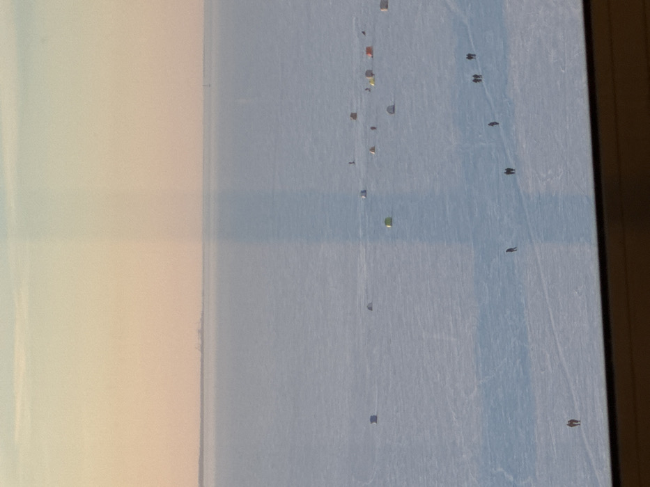This image presents a panoramic view of a vast, frozen body of water, identified as the Gulf of Finland adjacent to Saint Petersburg, Russia. The scene is observed from an elevated indoor position, with a dark horizontal window frame bordering the bottom and a prominent vertical light streak running down the center, indicating a window reflection or glare.
The sky displays a pale gradient, shifting from a light orange-pink at the horizon to a faded blue-white overhead, suggesting either early morning or late afternoon light conditions. The primary environment consists of a flat, expansive sheet of ice covered in white snow, exhibiting subtle textures and dark linear patterns indicative of foot traffic.
On the distant horizon to the left, a low-lying landmass with indistinct structural outlines is visible. To the far right horizon, a slender, dark, vertical structure, possibly a distant beacon or mast, is faintly discernible.
Scattered across the frozen surface are approximately 15 to 20 small, portable ice fishing tents. These tents vary in color, including shades of blue, green, orange, and muted earth tones, and are distributed across the mid-ground and background.
Numerous human figures are present, traversing the ice. In the lower left, two individuals walk together. Farther into the scene, a solitary figure walks rightward, while to their right, another pair walks in the same general direction. Towards the lower right, a group of three individuals walks, followed by an additional pair. Smaller, less distinct figures are also observed near several of the ice fishing tents, implying active participation in ice fishing. The overall scene depicts widespread ice fishing activity on the frozen gulf.