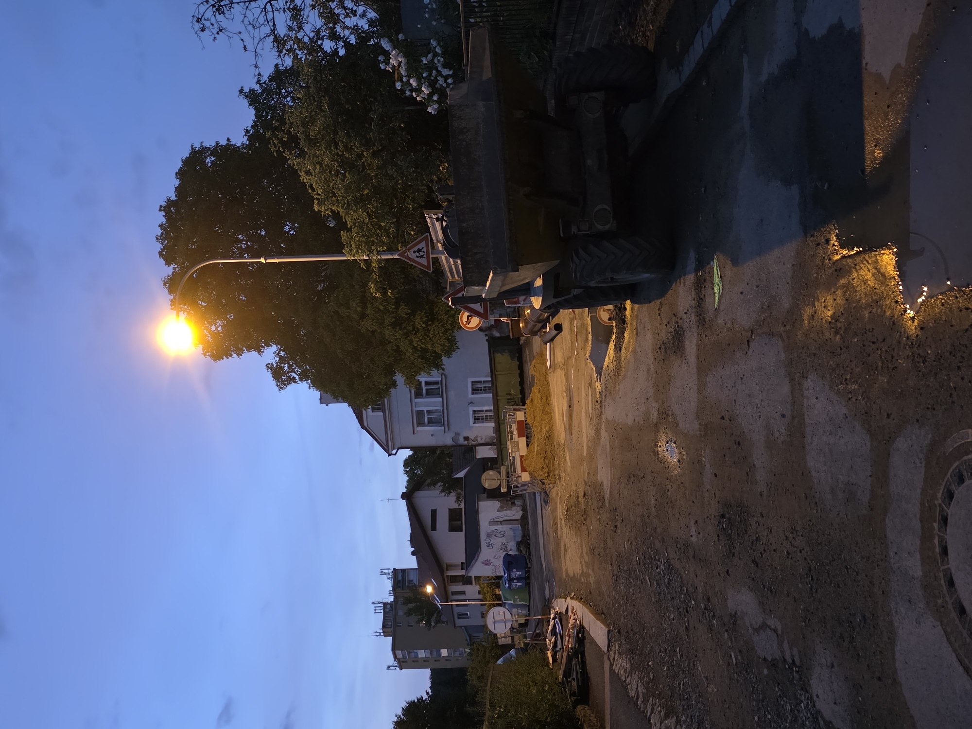 The image depicts an outdoor scene at dusk or dawn in an urban or suburban area, possibly undergoing construction or maintenance. A street lamp illuminates a partially paved road and a construction site. In the foreground, the ground is a mix of dirt, gravel, and puddles, suggesting recent rain or ongoing work. A large, yellow industrial dumper truck is parked on the right side of the road, its tires and rear end clearly visible. Various construction barriers, sand piles, and what appear to be discarded materials are scattered around the area. In the background, buildings of different sizes and styles can be seen, including what looks like an apartment block and older houses. There are trees and foliage, with white flowers blooming on a bush to the right. Several blue and green recycling bins are also visible near one of the buildings. Traffic signs, including a pedestrian crossing warning sign and a directional sign, are present. The sky is a deep blue, transitioning from lighter shades near the horizon, indicating twilight. No people are visible in the image.