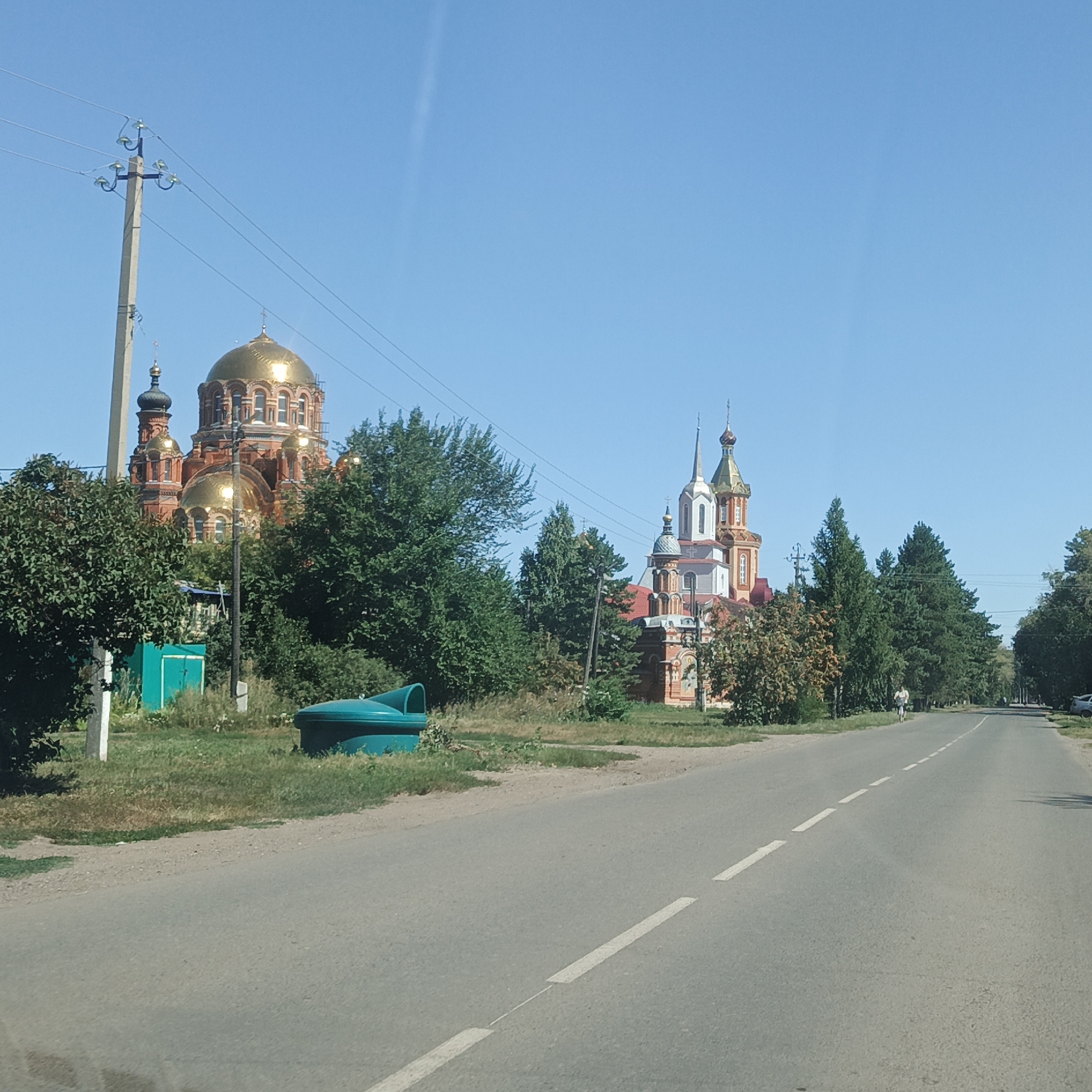 The image depicts a street scene in Saraktash, Russia. On the left side of the road, a prominent red brick church with golden domes is visible, partially obscured by lush green trees. To the left of the church, a utility pole with multiple wires stands tall against a clear blue sky. A bright green bin is positioned on the grassy verge next to the road. The road itself is paved with white dashed lines marking the lanes. In the distance, a person is seen cycling on the road, and a car is parked on the right side. The environment appears to be a sunny day with clear skies, suggesting a typical summer or early autumn scene in a rural or semi-urban setting.