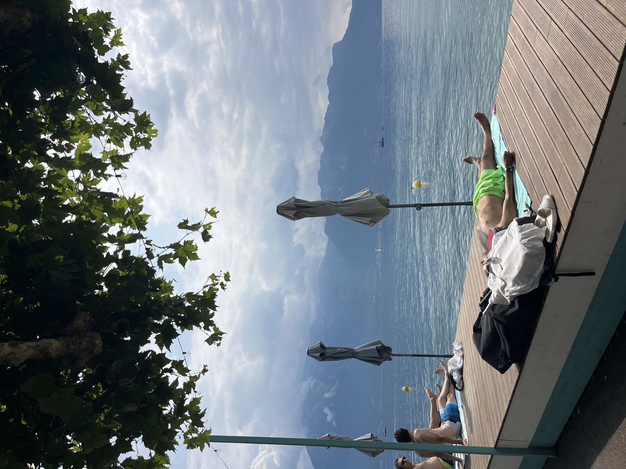 The image depicts a sunny day at the waterfront in La Tour-de-Peilz, Switzerland. In the foreground, a wooden deck area is set up for relaxation. Two men are visible on the deck. One man, wearing blue swim trunks, is sitting and looking towards the water. Another man, lying down and wearing green swim trunks, is sunbathing. Next to him on the deck are a black bag and some white clothing. Several closed gray umbrellas are spaced along the edge of the deck, suggesting this is a designated area for beachgoers. The calm blue water of a lake stretches out to the horizon, where a range of hazy, blue mountains forms a dramatic backdrop. The sky is partly cloudy, with bright patches of sunlight indicating fair weather. A sailboat can be seen in the distance on the water, adding a touch of activity to the serene landscape. The overall scene conveys a relaxed atmosphere of leisure and enjoyment by the water.