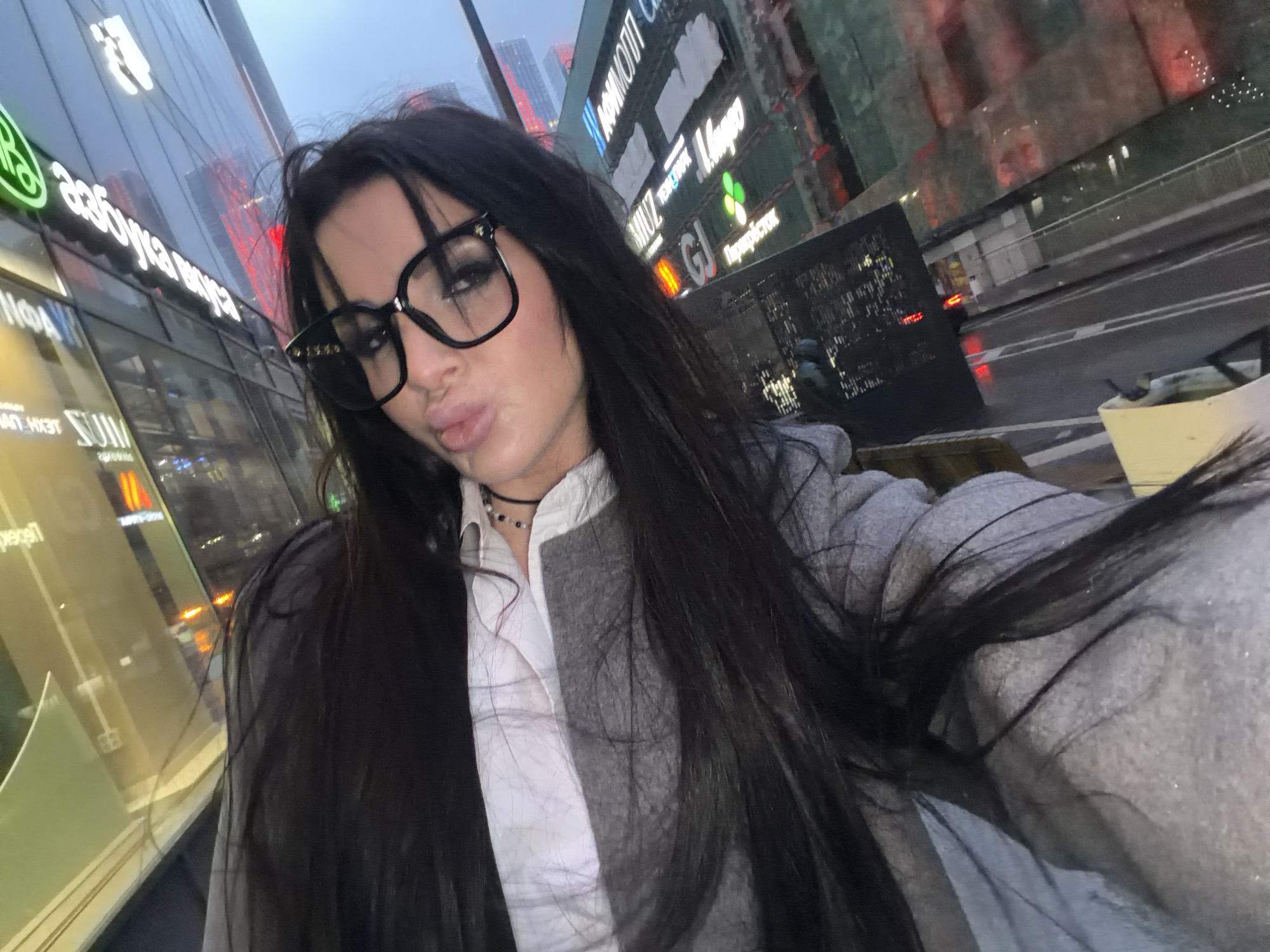 A medium-shot selfie shows a woman with long black hair, wearing large, dark-rimmed glasses, a white collared shirt, and a gray jacket, posing with pursed lips. The background suggests an urban environment, with visible buildings and signs in Cyrillic script, indicating a location in Russia, specifically the city of Khimki, based on the Cyrillic lettering on the buildings. A street with wet surfaces is visible on the right, suggesting it may have recently rained.