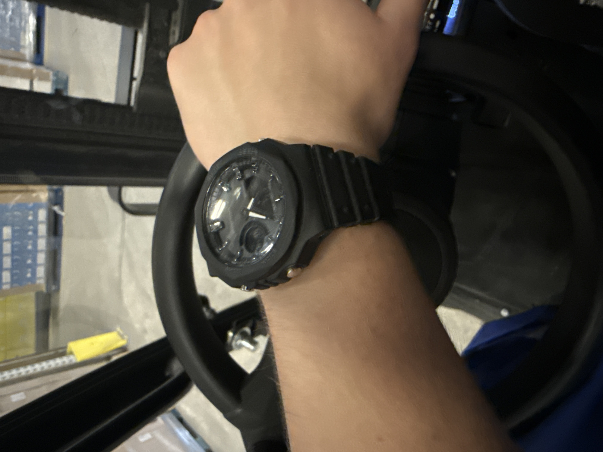 A first-person perspective shot shows a person's arm and wrist wearing a black digital watch. The person is seated in what appears to be a forklift, as indicated by the steering wheel. The background suggests an industrial or warehouse setting, with pallets of goods visible. The scene is lit with overhead lighting, common in such environments. The overall impression is that of someone operating machinery in a commercial or industrial facility.