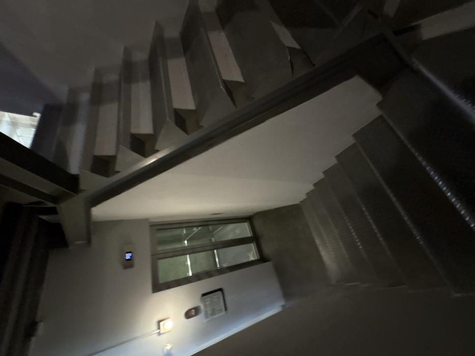 A low-angle shot depicts a stairwell. Metal stairs rise on both sides of the frame. A wall with a window is visible on the left, featuring an electronic device and some fixtures. The image exhibits a slightly tilted perspective.