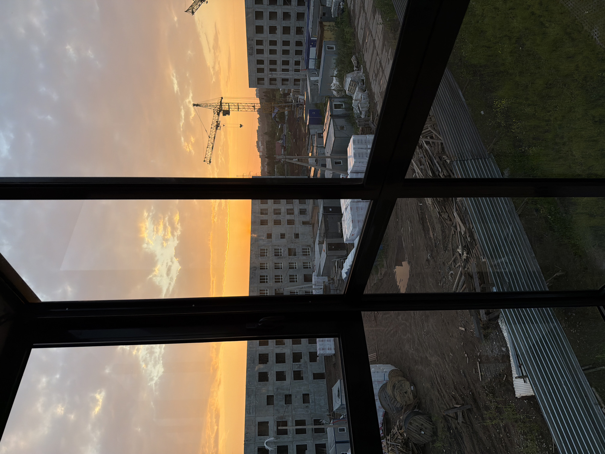This is a high-angle, full shot photograph taken from inside a building, looking out through a window with dark frames. The image captures a construction site at sunset. The sky is a vibrant mix of orange and yellow hues with scattered clouds. In the distance, two tower cranes are visible against the sunset. In the middle ground, the skeletal structures of unfinished apartment buildings are prominent, featuring rows of dark rectangular windows. Surrounding these buildings are various temporary structures, possibly portable offices or storage units, some of which are covered with white tarps. The ground appears to be dirt and gravel, with piles of construction materials, including large wooden spools and stacks of lumber. A corrugated metal fence runs along the foreground. The overall impression is one of ongoing urban development in the city of Froly, Russia, during the evening hours. No people are visible in the frame.