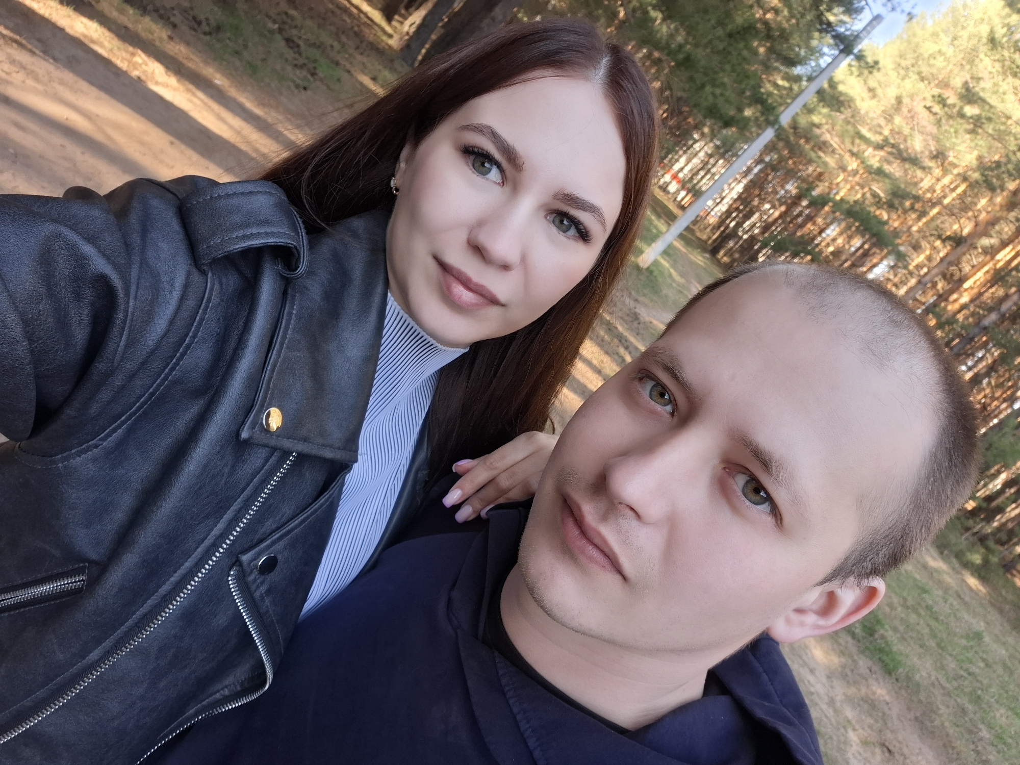 A selfie taken outdoors shows a woman and a man posing closely together. The woman, with brown hair, is wearing a black leather jacket over a light blue and white striped shirt. Her manicured hand rests on the man's shoulder. The man is wearing a navy blue hooded sweatshirt. The background shows a park or wooded area with tall trees, possibly taken in Chaikovsky, Russia. A lamp post is visible among the trees. The lighting suggests it is daytime.