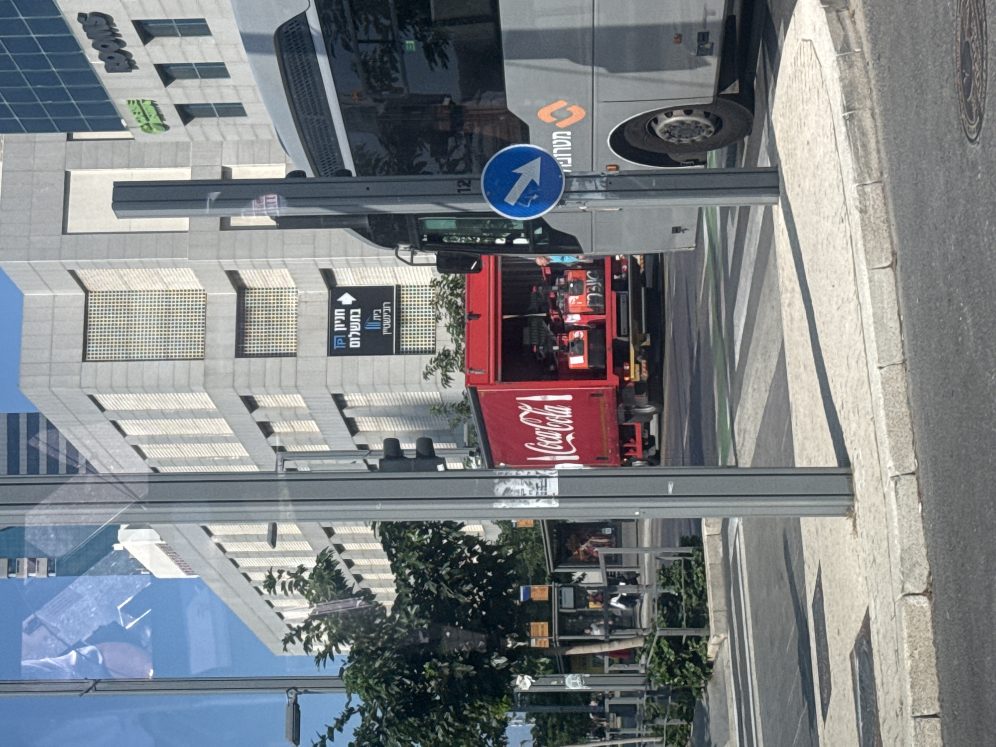 The image captures a street scene in Tel Aviv, Israel, with a focus on a Coca-Cola delivery truck and a bus. The truck, painted bright red with the iconic Coca-Cola logo, is partially visible on the left, its rear doors open, revealing stacked cases of bottles. To the right, a gray bus is parked, its front section and wheel clearly in view. A blue circular traffic sign with a white arrow pointing diagonally down and to the right is affixed to a gray pole in front of the bus. In the background, modern buildings with geometric facades line the street, one bearing the signage "ROOMS" in green. A traffic light is visible on a pole in the foreground, partially obscured. The scene appears to be at a street intersection, with pedestrian crossings and sidewalks evident. The lighting suggests it is daytime, with shadows cast on the ground.