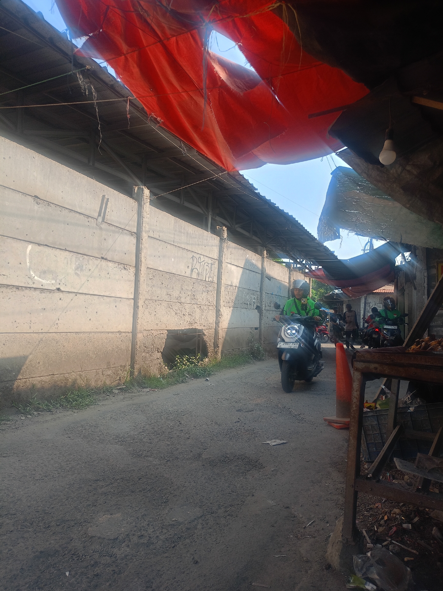 The image depicts a narrow, unpaved road in Bojong Gede, Indonesia. A man wearing a green jacket and helmet is riding a scooter on the right side of the road, heading towards the viewer. Another scooter with a rider wearing a similar green jacket is visible in the background. The road is flanked by a tall, textured concrete wall on the left and a makeshift structure with a corrugated metal roof and a torn red tarp overhead on the right. The environment appears to be somewhat rundown, with debris and scattered litter visible along the roadside. The lighting suggests it is daytime, with sunlight filtering through the torn tarp.