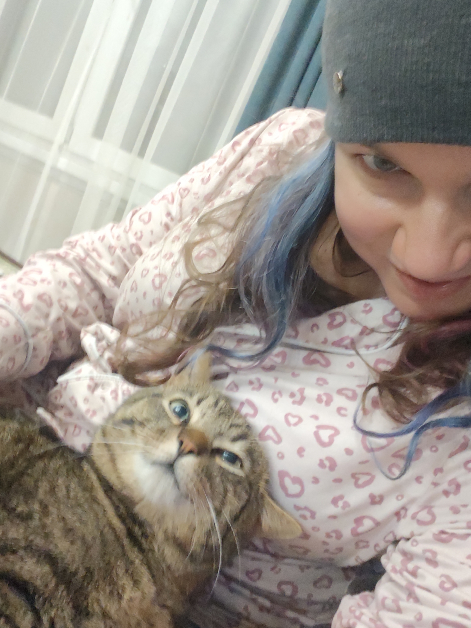 A selfie depicts a person wearing a gray beanie and a pink and white heart-patterned top, cradling a brown tabby cat. The person has blue streaks in their hair. The location is suggested to be in the vicinity of Свистуха, кузяевский С/о, Russia, based on provided location data (not visible in the image). The background shows light-colored curtains. The cat is looking upwards with a slight tilt of its head. The person is looking down at the camera.