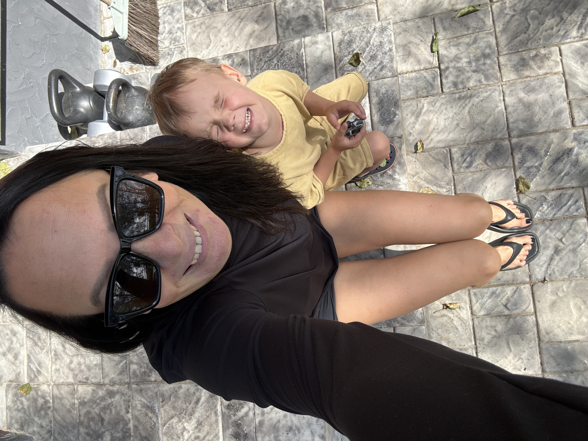A high-angle, full-body selfie shows a woman and a young child outdoors. The woman, positioned in the upper left of the frame, is wearing sunglasses and a black t-shirt. She is smiling, and her long, dark hair is visible. The child, in the center right, is wearing a yellow shirt and shorts, with eyes closed and a wide smile. The child is holding a small toy. They are both standing on a paved patio with a textured, stone-like pattern. Fallen leaves are scattered on the ground. In the background, to the right of the child, a set of kettlebells and a broom leaning against a textured wall are visible. The scene appears to be a casual outdoor setting, potentially a backyard or patio area. The location in Chaikovsky, Russia is not discernible from the visual information provided in the image.