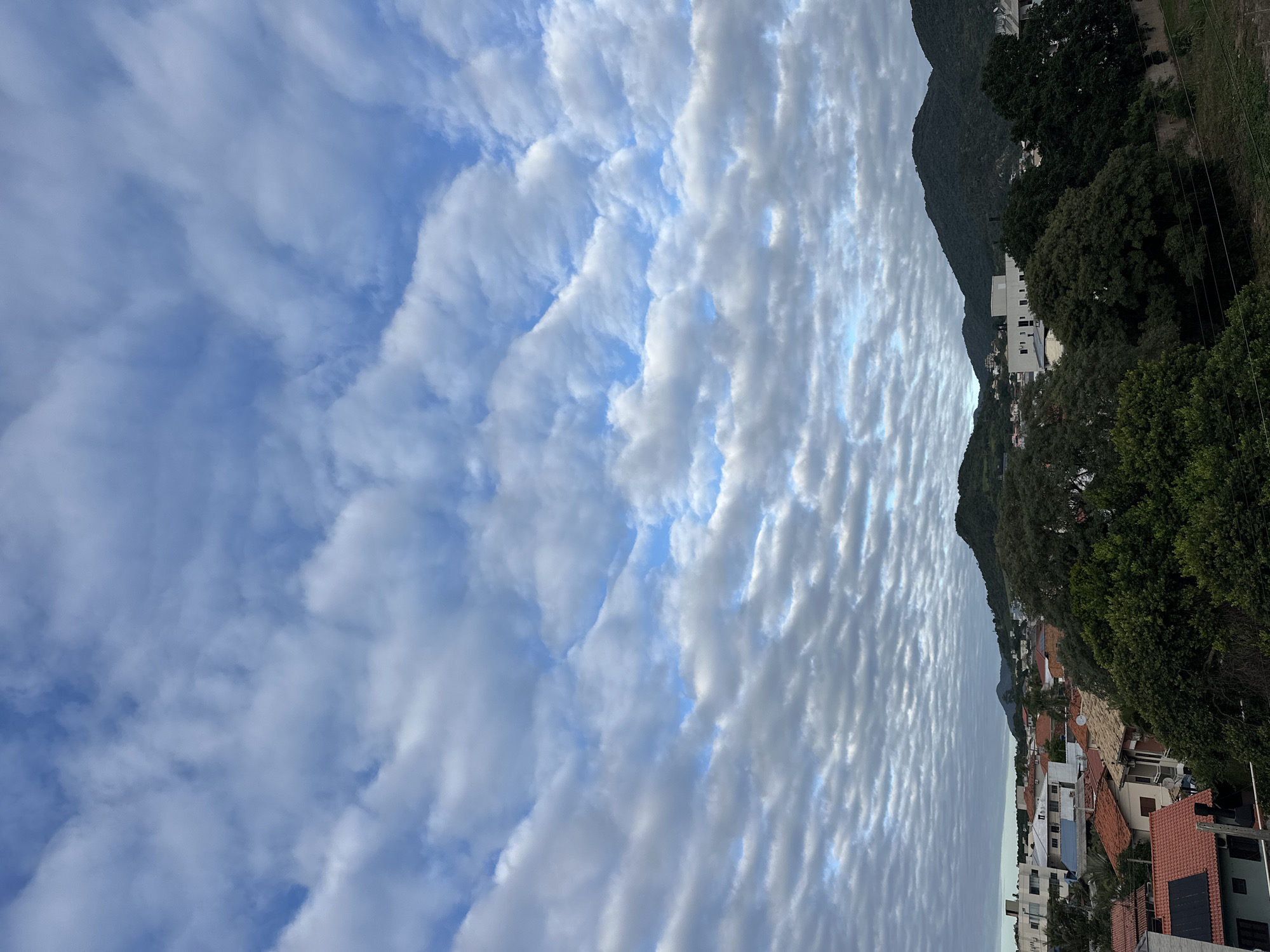 The image shows a view in Florianópolis, Brazil. The sky is filled with altocumulus clouds. On the right side of the frame, there are hills covered with dense vegetation and some residential buildings with red-tiled roofs. The angle of the shot suggests it was taken from an elevated position. There are no people visible.