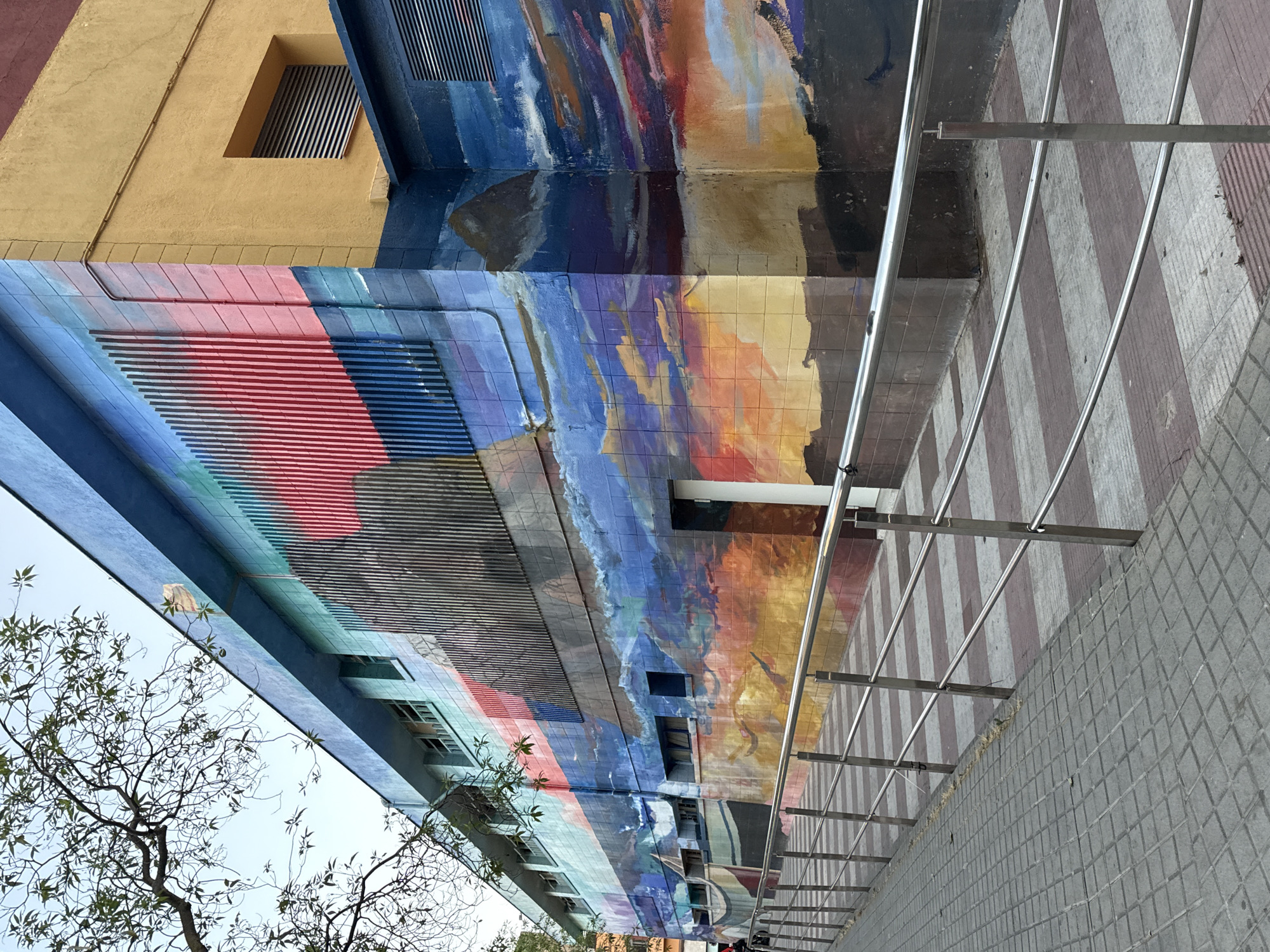 The image shows a colorful mural painted on the exterior of a building in Blanes, Spain. The mural depicts a vibrant sky with clouds, possibly a sunset or sunrise, with shades of orange, yellow, blue, and purple. There are also elements that suggest boats and landforms, giving a coastal or maritime feel. The building itself appears to be multi-story with windows. A metal ramp with handrails leads up to an entrance on the right side of the mural. The ground in front of the ramp is paved with square tiles, and a portion of a tree with green leaves is visible in the upper left corner. The mural extends along the side of the building, wrapping around a corner.