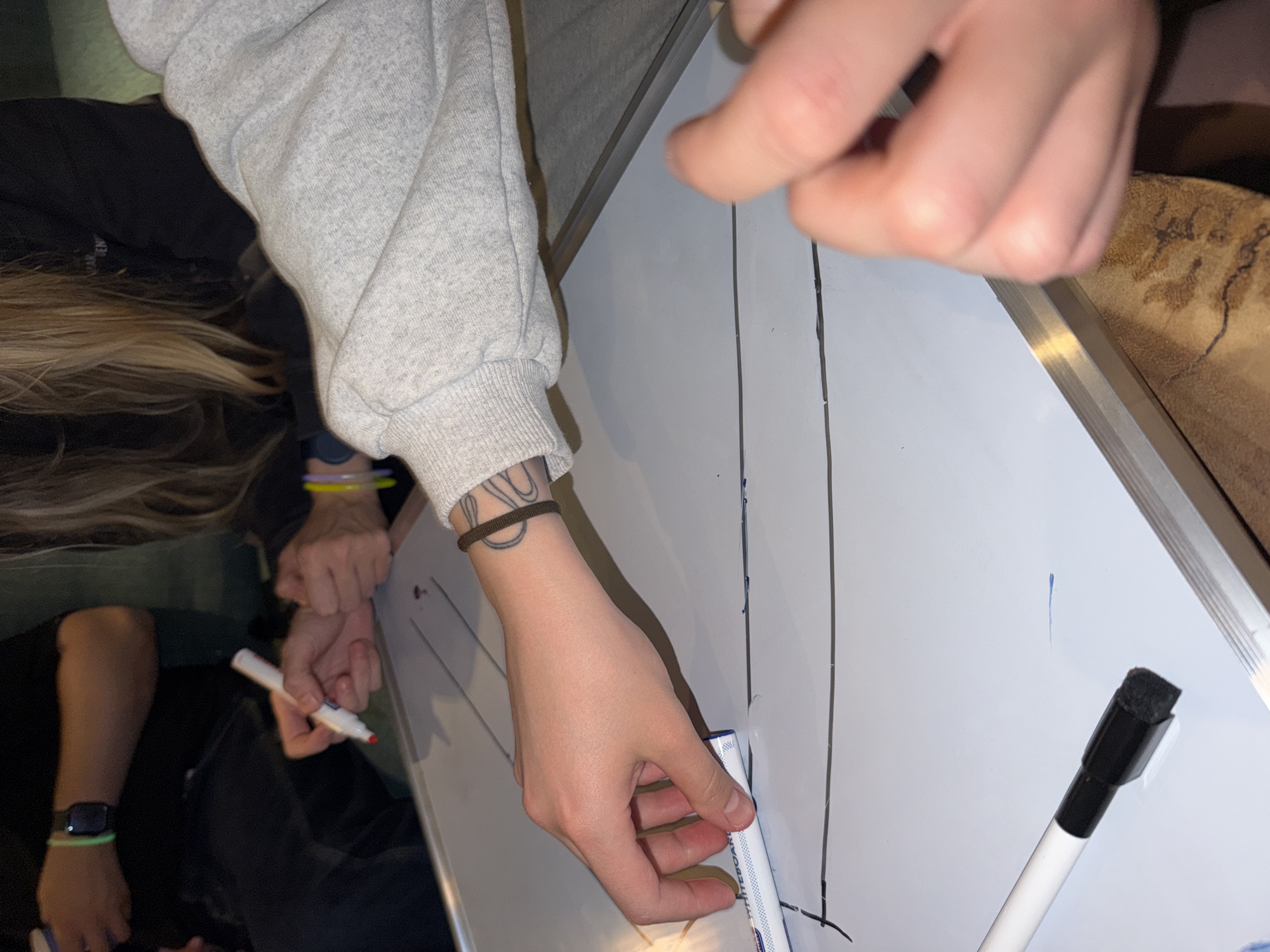 The image shows a close-up view of a whiteboard with black lines drawn on it, suggesting some form of diagram or planning is taking place. One person's hand, with a dark elastic bracelet on their wrist and tattoos on their forearm, is holding a white whiteboard marker with a red tip. Another person's hand is visible in the lower right corner of the frame, also near the whiteboard. In the background, other individuals are partially visible, one wearing a black long-sleeved shirt with a smartwatch on their left wrist and a green wristband, and another with blonde hair and colorful wristbands. The overall environment appears to be indoors, possibly a meeting room or study area, with a focus on the activity of writing on the whiteboard. The city of Heijen in the Netherlands is not depicted or implied in this image.