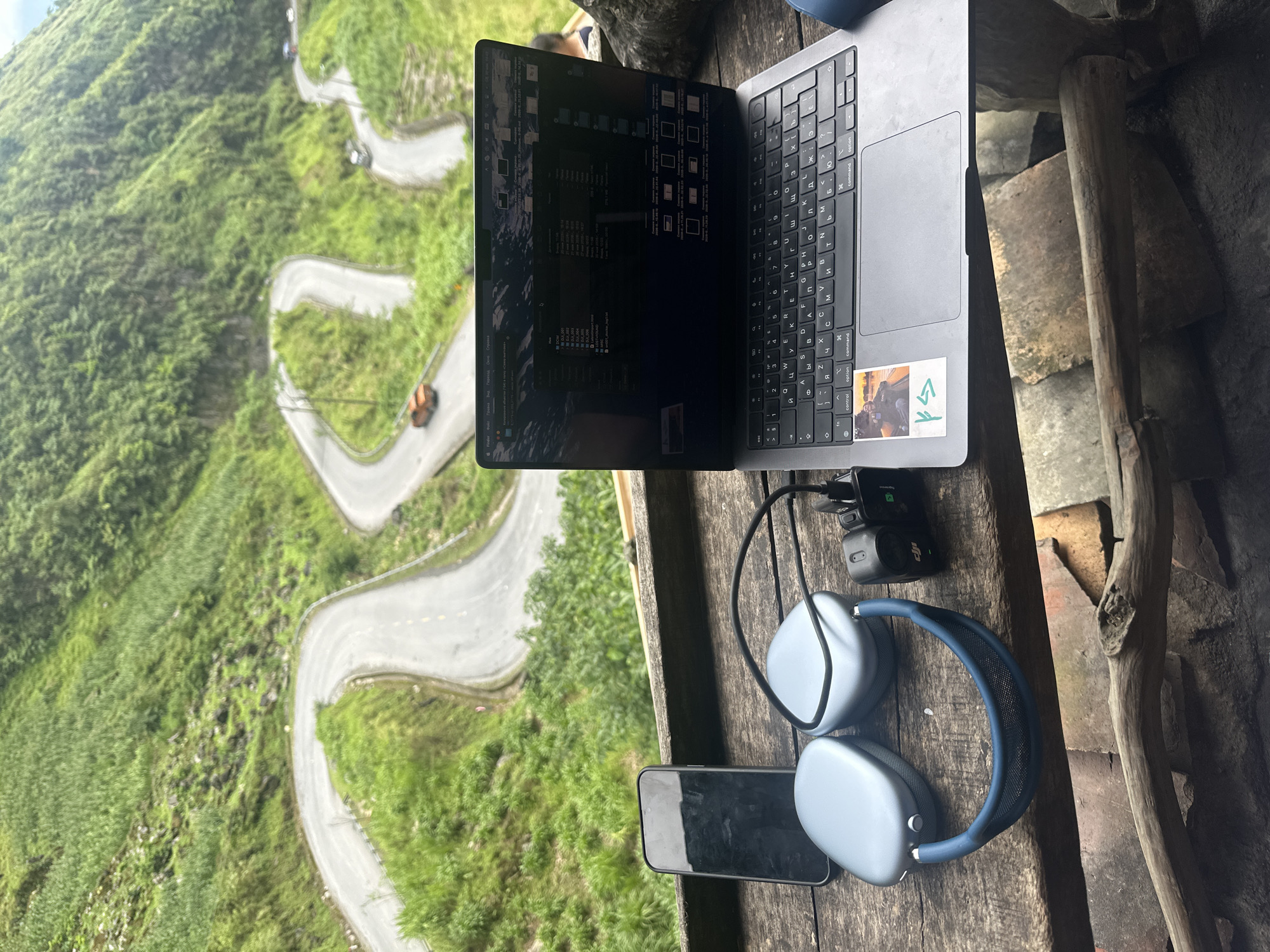 Here is a detailed description of the image:
**Scene:**
The image captures a viewpoint from what appears to be a rustic, wooden table or platform overlooking a striking landscape. It's a workstation set up in an unconventional setting, with a blend of modern technology and natural scenery.
**Objects:**
* **Laptop:** A modern laptop, seemingly a Macbook, is the central focus. Its screen shows a display of code or file directories, suggesting work in progress.
* **Headphones:** A pair of high-end, noise-canceling headphones (AirPods Max).
* **Smartphone:** A smartphone lies near the headphones.
* **Charger:** A small charger connected to the laptop, indicating a power source.
* **Wooden Structure:** The scene is framed by a wooden structure.
* **Sticker:** A sticker is attached to the keyboard section of the laptop.
**Environment:**
* **Road:** A winding road with multiple hairpin turns. Vehicles can be seen on the road.
* **Vegetation:** The hillsides are covered in lush greenery, implying a subtropical or mountainous region.
* **City of Yên Minh Commune, Vietnam:**
* **Geography:** The winding road indicates the presence of elevated mountains.
**Actions:**
The photo suggests a person is working remotely, combining the comforts of technology with the beauty of the surrounding landscape.