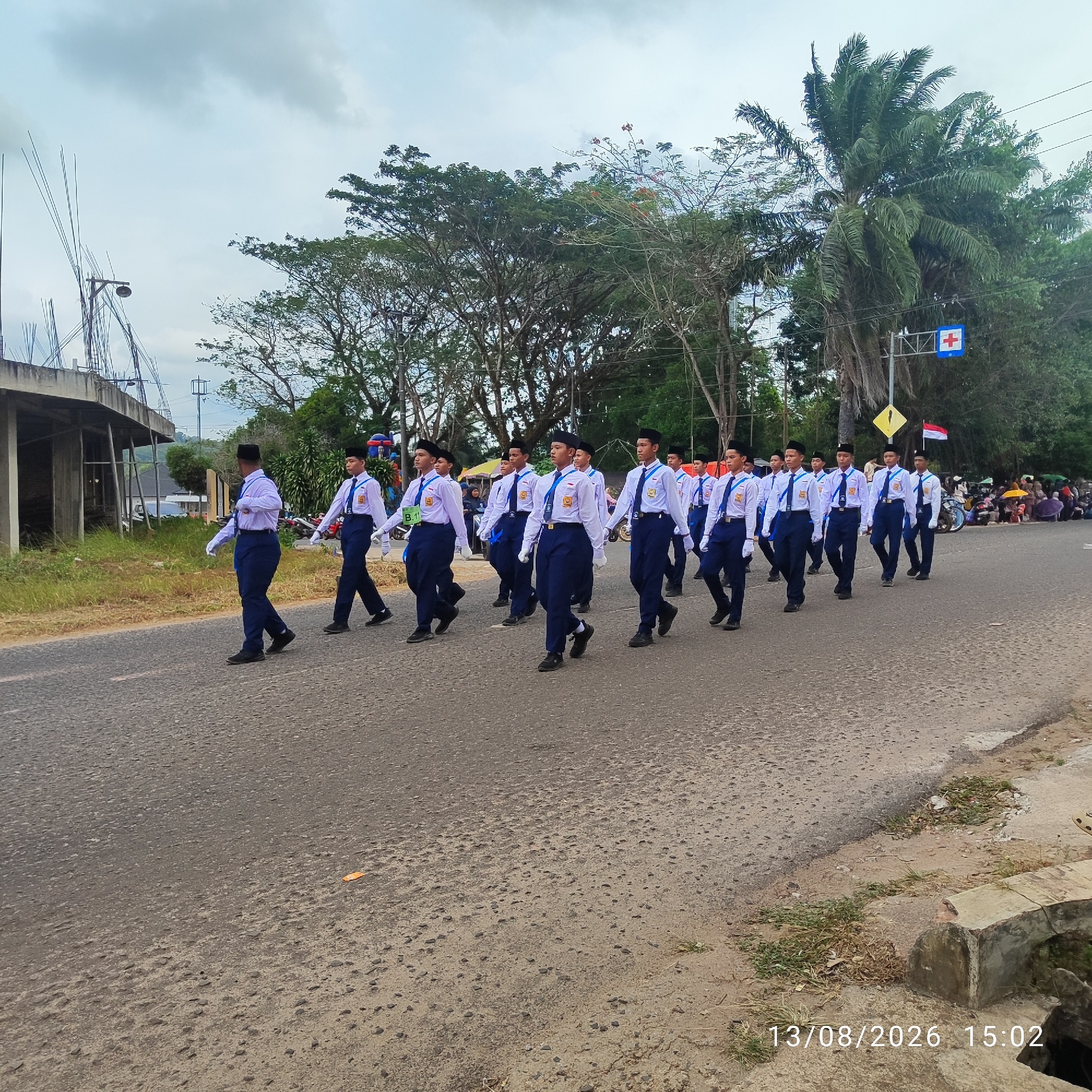 A group of uniformed students are marching in formation along a paved road in Buding, Indonesia. The students, wearing white shirts, dark blue pants, black hats, and white gloves, are moving from left to right in the frame. They appear to be participating in a parade or official procession. Behind them, on the right side of the road, are spectators seated under umbrellas and a few motorcycles parked. A traffic sign with a red cross symbol is visible on a pole. To the left of the road, there is an unfinished building structure with exposed rebar and a concrete foundation, surrounded by overgrown grass. The sky is overcast with clouds. The timestamp "13/08/2026 15:02" is overlaid on the bottom right of the image.