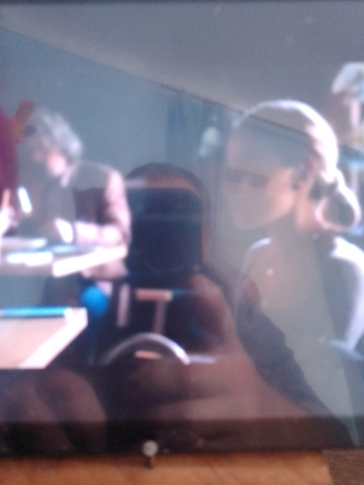 The image shows a dimly lit, slightly blurry interior scene. Several people are visible, mostly out of focus. In the foreground, on the left, a person is partially visible with a pinkish hue on their left arm. To their right, a dark, upright object that resembles a monitor or screen is present. In the mid-ground, a person with light-colored hair styled in a bun is looking to the right, and their face is illuminated. To the left of this person, another individual's head and shoulder are visible. The background is indistinct, with some lighter areas that could be walls or other surfaces. There are horizontal surfaces in the scene that appear to be tables or desks, with some lighter-colored objects on them. The overall impression is of an indoor gathering or workspace. The image does not contain any discernible information that would indicate a specific location in the city of Kotiv, Ukraine.