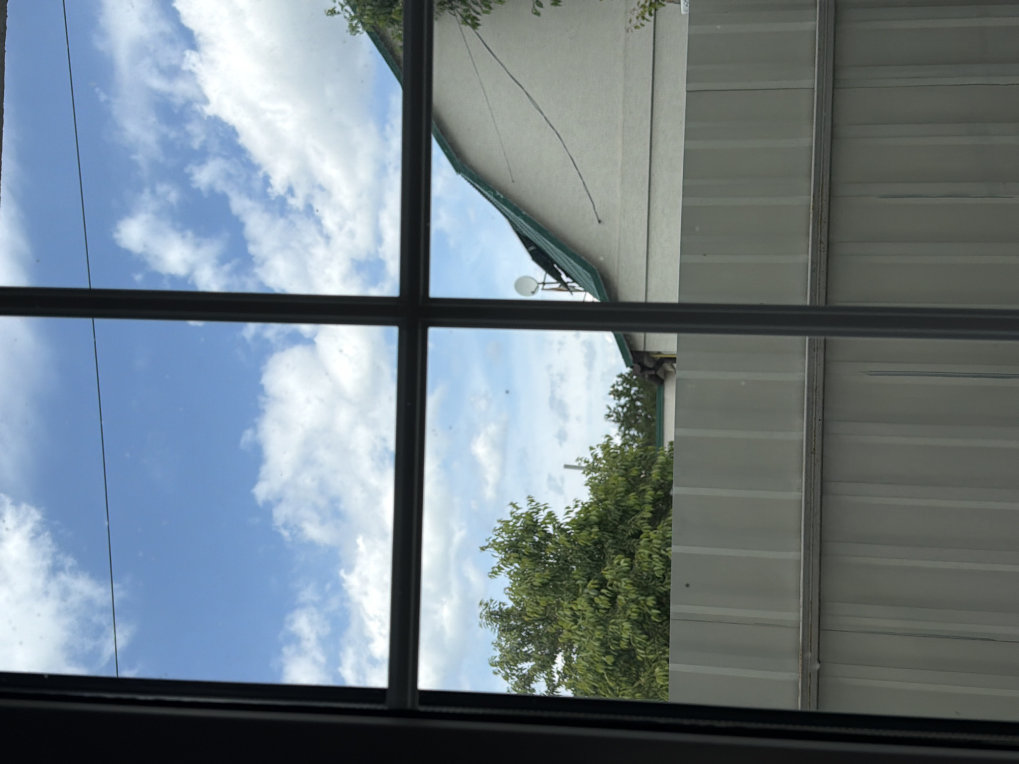 A view through a window shows a cloudy blue sky with a thin wire stretching across the frame. Below the sky, a green-roofed building is visible, with a white facade and a satellite dish on its roof. Trees with dense green foliage are to the left of the building. A gray corrugated metal fence runs horizontally across the lower portion of the image, dividing the scene. The image appears to be taken from a residential area, possibly in Bila Tserkva, Ukraine, given the typical architecture and fencing.