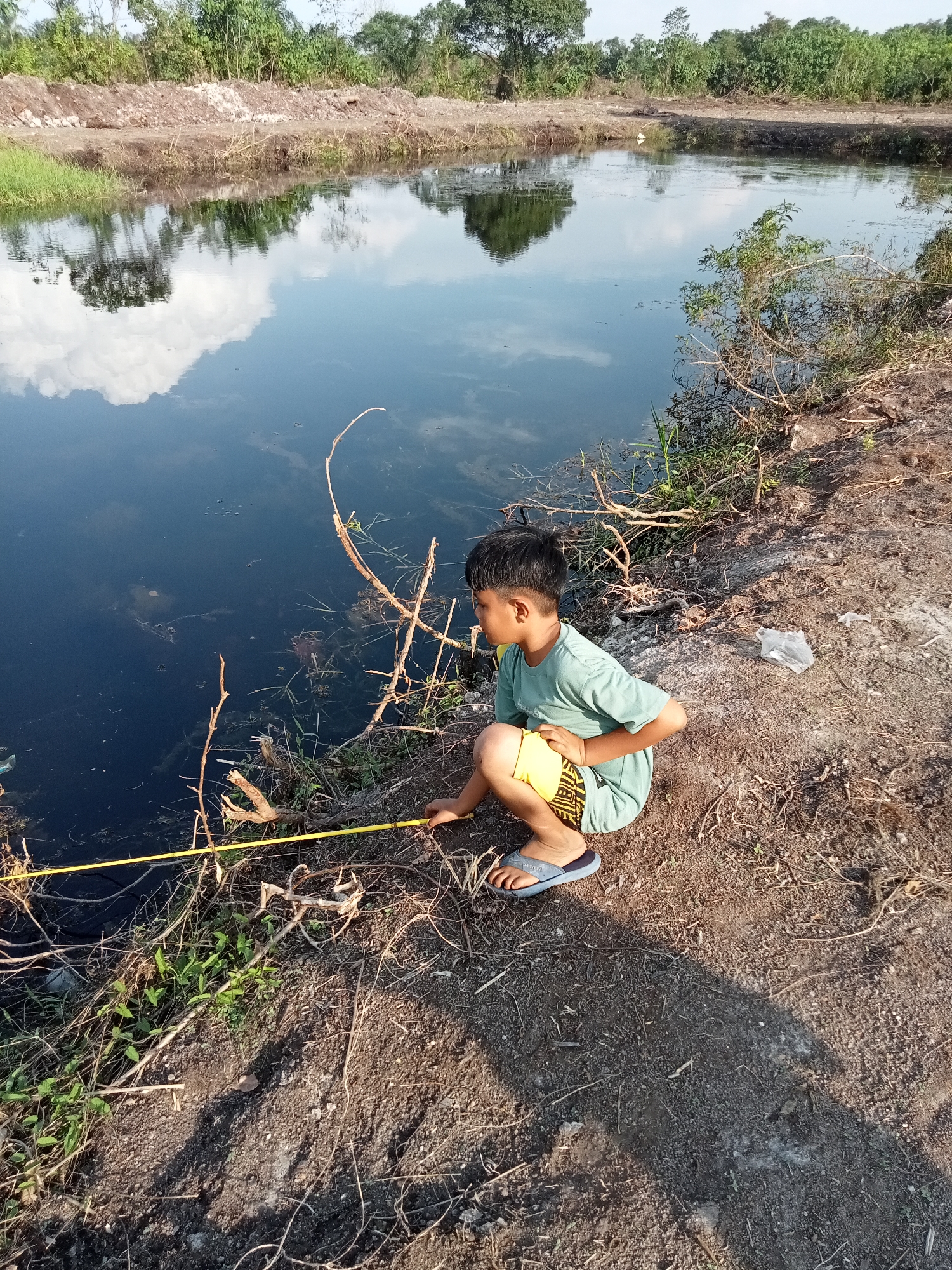 In Teluk Kenidai, Indonesia, a young boy is crouched beside a dark, still body of water. He is wearing a light teal t-shirt and yellow shorts with a black patterned waistband. His feet are clad in blue flip-flops. In his hands, he holds a yellow measuring tape which extends out towards the water. The boy appears to be focused on something in or near the water. The surrounding environment is natural and somewhat unkempt, with dry grass and fallen branches along the water's edge. The water reflects the sky and surrounding trees, indicating a calm day. The ground is a mixture of dirt and small stones. In the background, there are trees and a disturbed area of earth, suggesting ongoing construction or land modification. A crumpled white plastic bag is visible on the ground behind the boy. The scene appears to be an outdoor, rural setting.