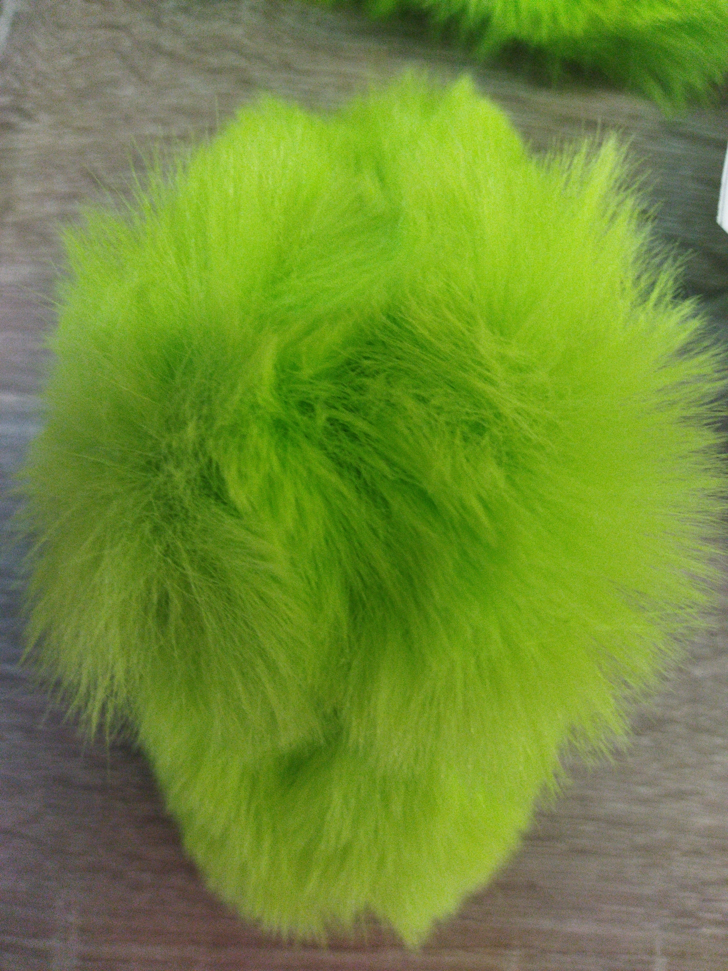 A close-up, top-down shot of a bright green, fluffy pom-pom rests on a wooden surface. The pom-pom is round and appears to be made of faux fur, with individual strands of varying lengths creating a textured, voluminous appearance. The wooden surface is visible in the background, appearing to be made of planks with a wood grain texture. The lighting suggests an indoor setting, though no specific location or environmental context beyond the immediate objects is discernible.