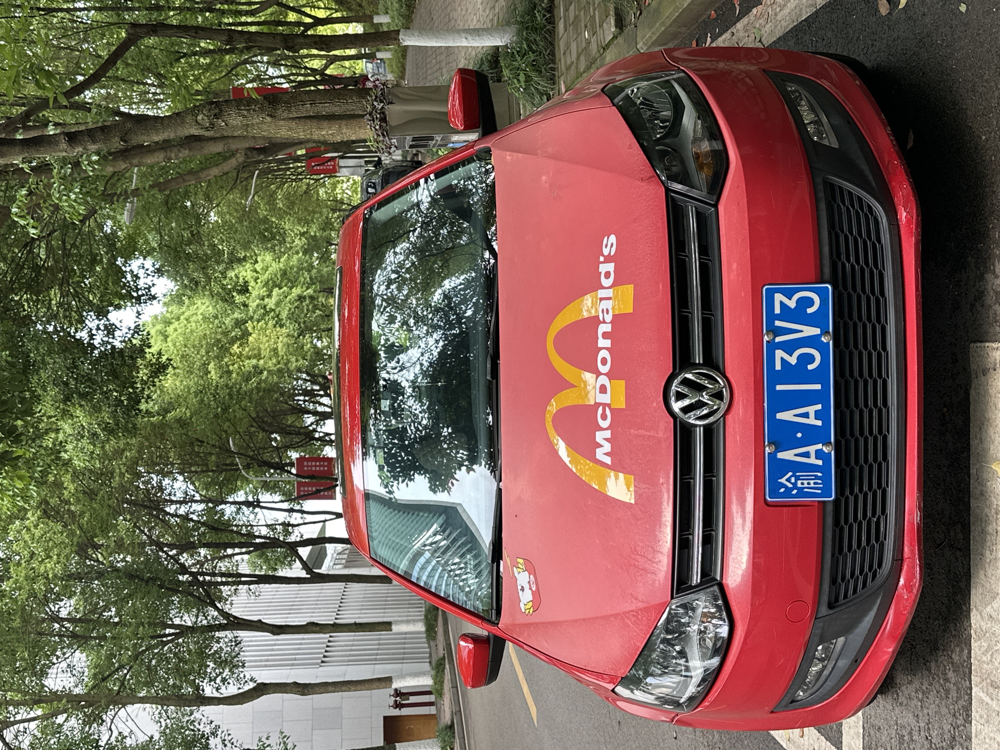 A red Volkswagen is parked on the side of a road. A McDonald's logo is prominently displayed on the hood of the car. The car has a blue license plate with the characters 渝A·A13V3. Based on the license plate prefix "渝A," the location is identified as the city of Jiangjin, China. The street is lined with trees and a building is visible in the background.