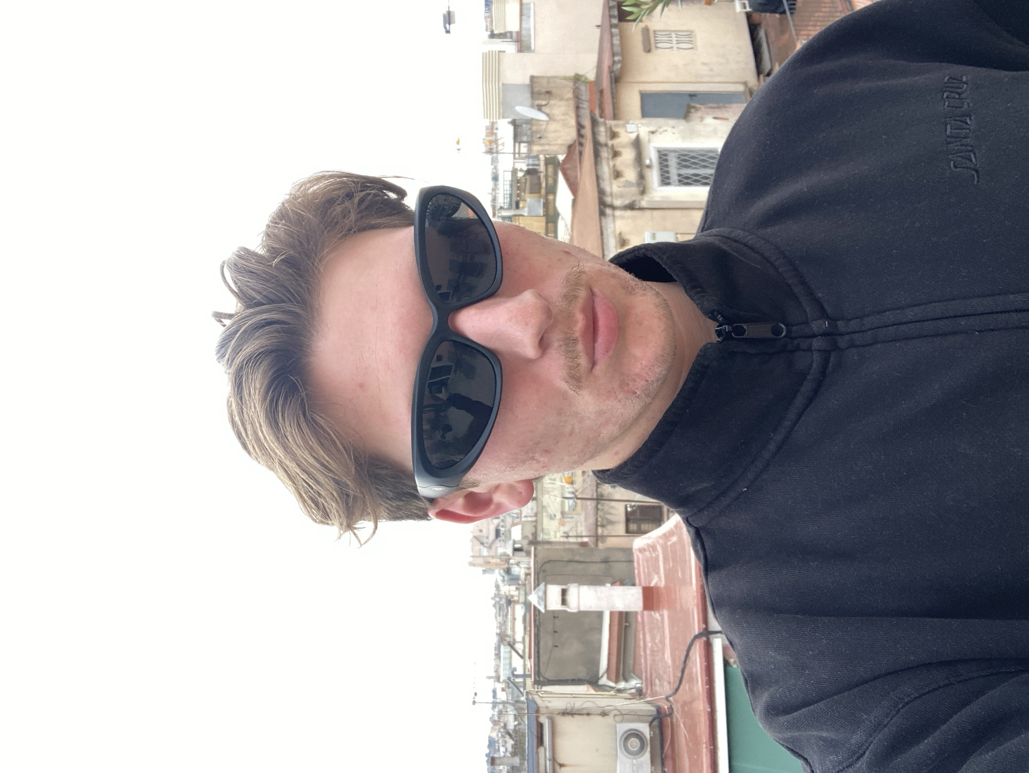 A selfie shot of a fair-skinned person wearing sunglasses and a dark-colored zip-up jacket with "SANTA CRUZ" printed on the shoulder. The person has short, light brown hair and a light mustache. They are positioned with a medium shot facing the camera. The background is a cityscape, likely taken from a rooftop. The architectural style of the buildings suggests the city is Barcelona, Spain. Elements in the background include rooftops, antennas, windows, and various building structures. The sky is overcast and grey.