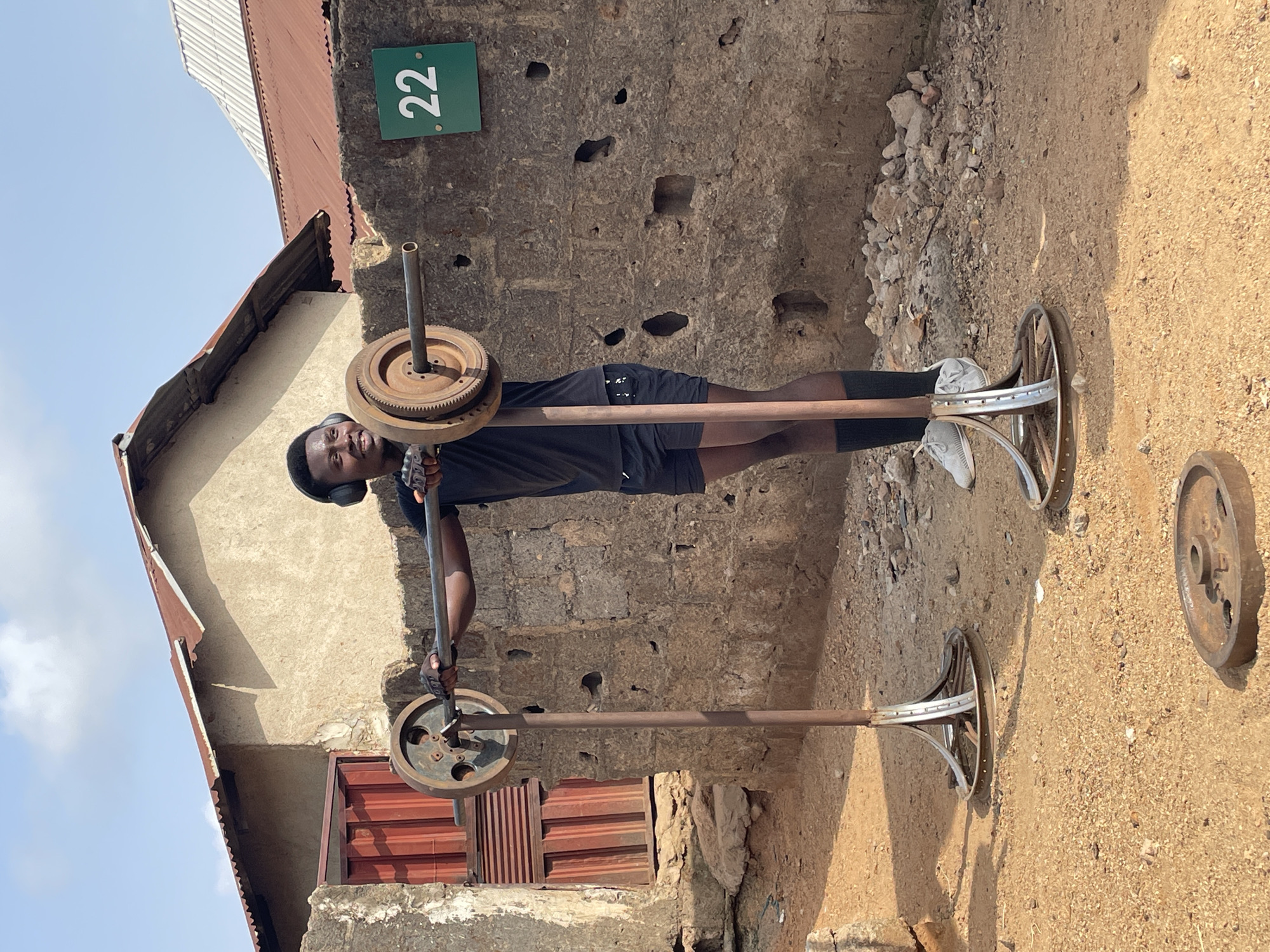 The image captures a young man engaged in weightlifting outdoors in Akwanga, Nigeria, under a bright, clear sky. He stands center-frame, looking directly at the camera with a determined expression, holding a homemade barbell at waist height. He is dressed in a black t-shirt, black shorts, black socks, white sneakers, and dark headphones, with weightlifting gloves on his hands.
The weightlifting equipment is notably crafted from repurposed materials. The barbell itself is made from a thick metal pipe or rebar, with large, circular metal components, possibly industrial gears or flywheels, serving as weight plates on either end. The barbell rests on two upright stands ingeniously fashioned from bicycle wheel rims. Another identical metal weight plate lies on the sandy-brown ground to the right of the man.
The setting is an unpaved outdoor area with sandy soil. Behind the man is a rugged wall constructed from rough stone or concrete, displaying several indentations. Above the wall, part of a building with a reddish-brown corrugated metal roof is visible against the sky. A small, rectangular green sign with the white numbers "22" is affixed to the wall to the left of the man's head. The bright daylight suggests it is midday or early afternoon. The scene highlights resourcefulness in fitness.