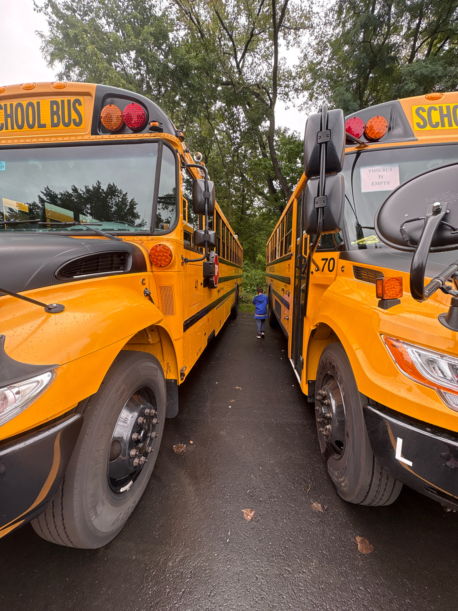 Two yellow school buses are parked side-by-side on a wet, asphalt surface outdoors, likely a school parking lot. The bus on the left is closer to the viewer, and its front left tire is prominently featured. The bus on the right is slightly further away, and its front right tire is visible. Both buses have "SCHOOL BUS" printed in yellow on their upper front sections. A small child in a blue jacket is walking between the two buses, away from the camera. The environment is overcast with trees in the background. The location is identified as Newington, United States of America.